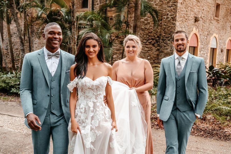 Wedding party: bride in white floral gown walks with attendants in front of stone building.