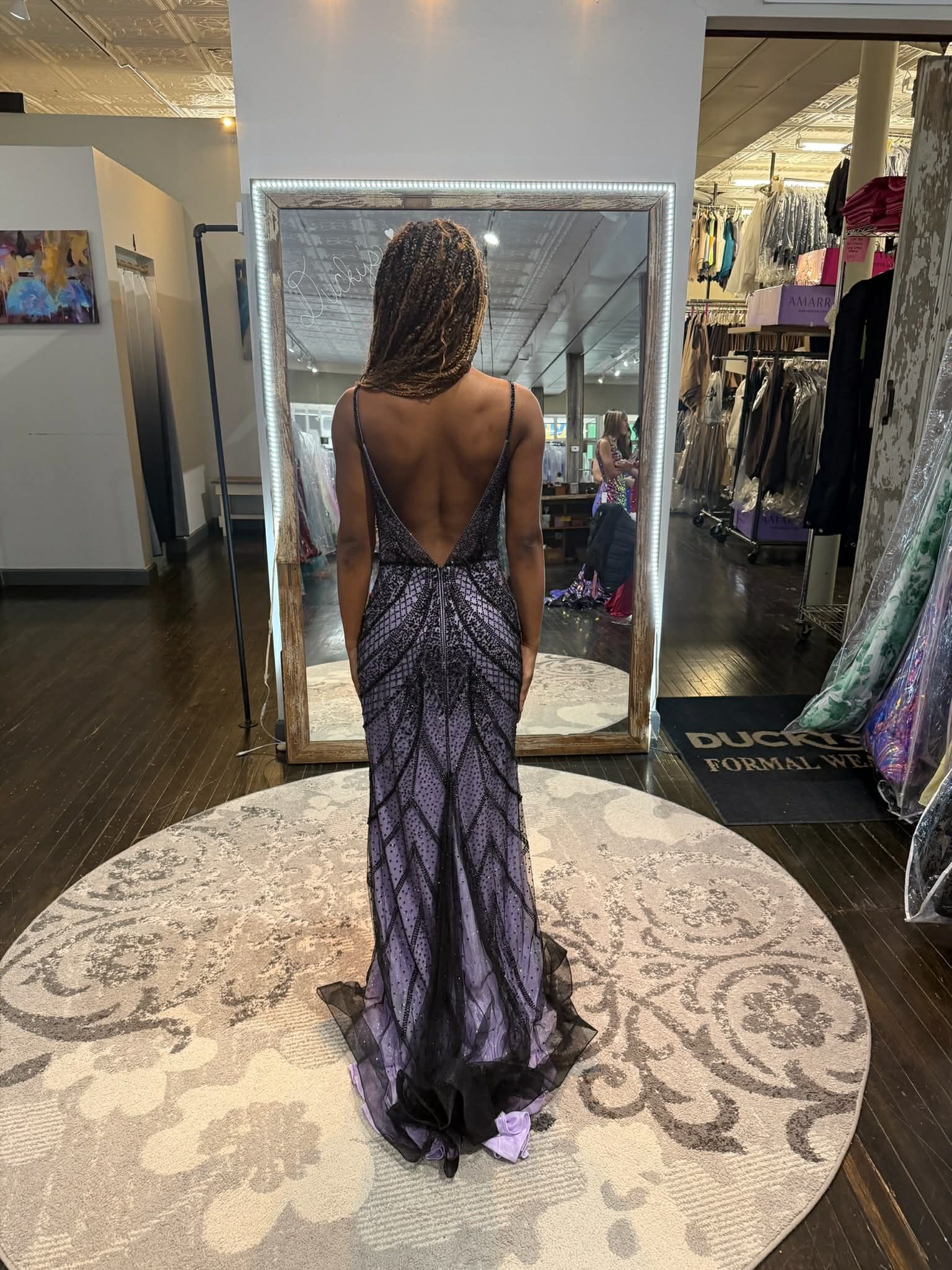 A person stands on a circular rug in a boutique, wearing a backless purple and black patterned gown in front of a mirror.