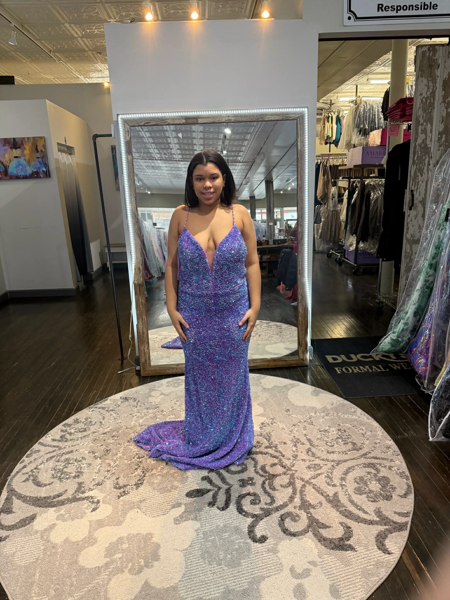 A person stands on a circular rug in a store, wearing a floor-length, shimmering purple sequined gown with a V-neckline.