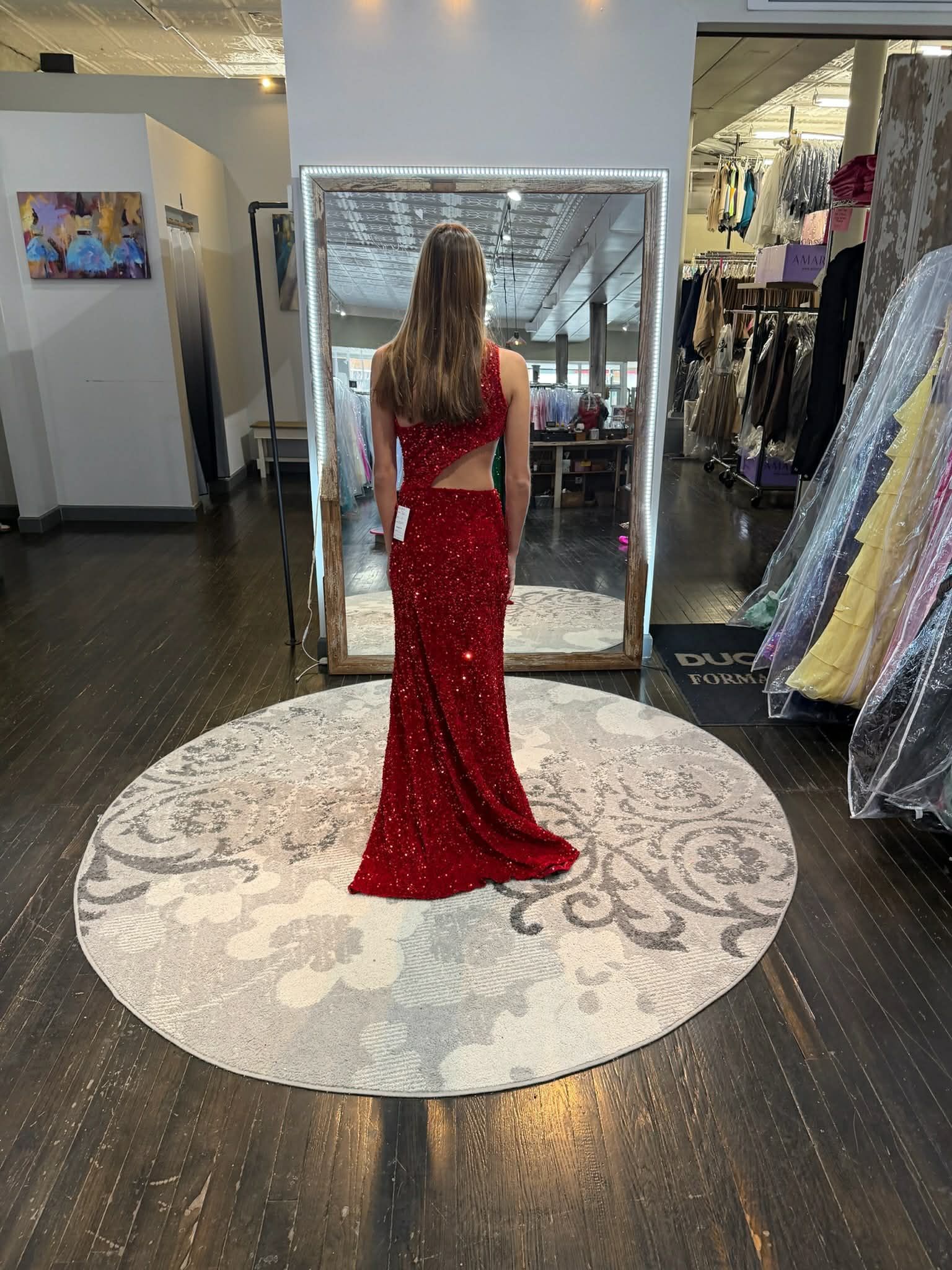 A person wearing a sparkly red floor-length gown stands on a circular rug in front of a mirror in a clothing store.