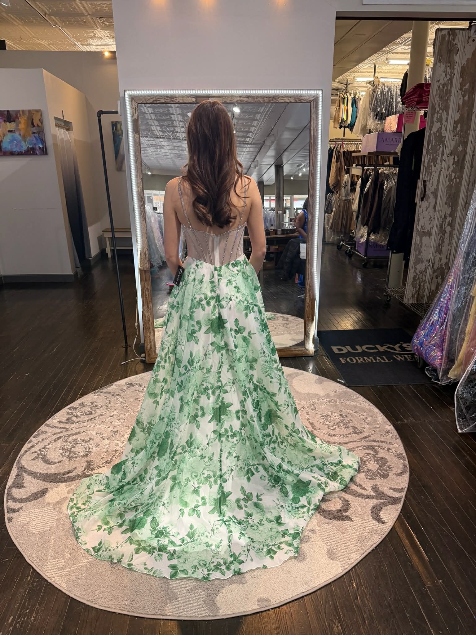 A person stands in a clothing store mirror wearing a white, floor-length dress with a green floral print and a train.