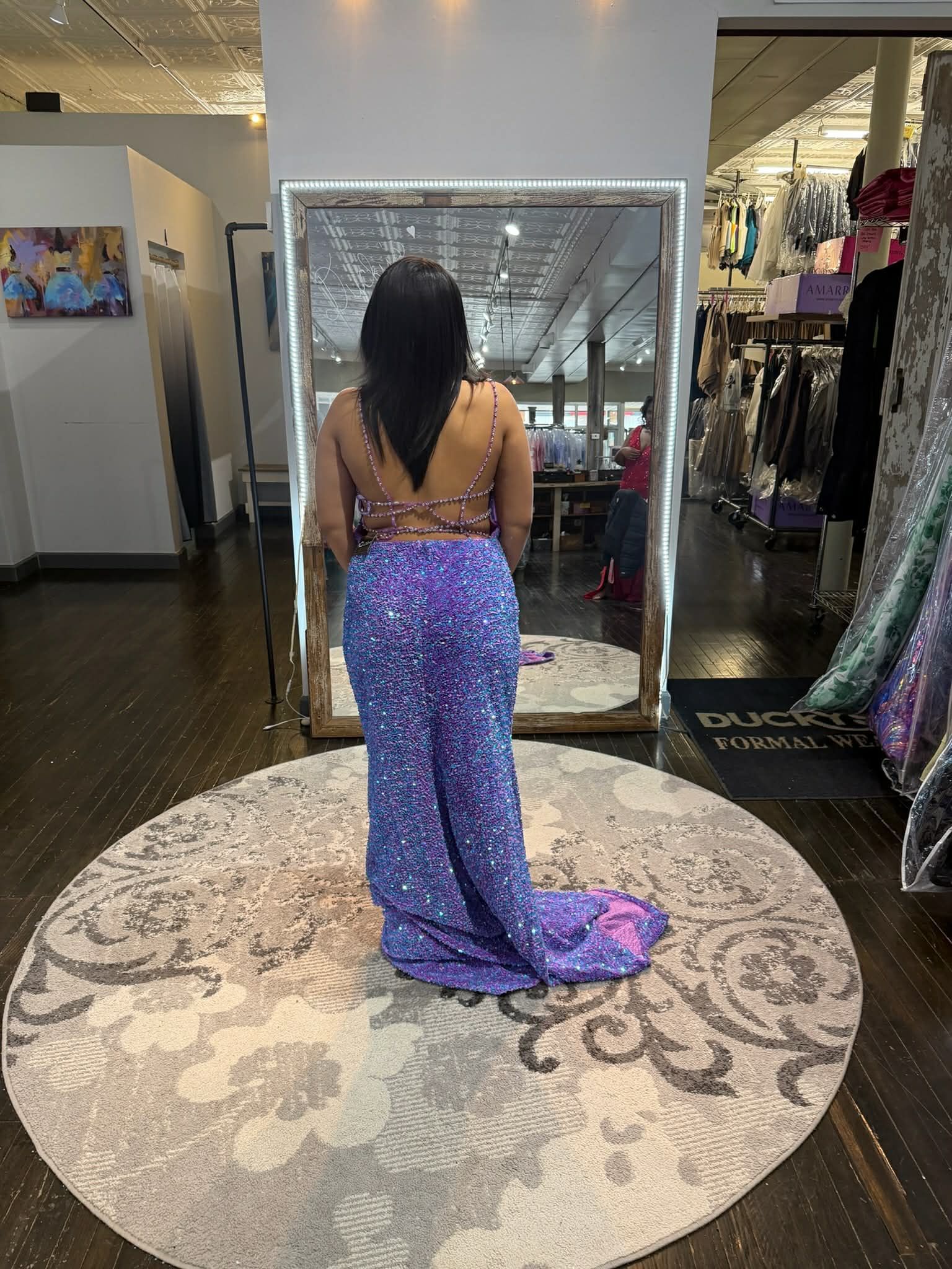 A person wearing a backless, sequined purple gown stands on a round rug, facing a large mirror in a clothing store.