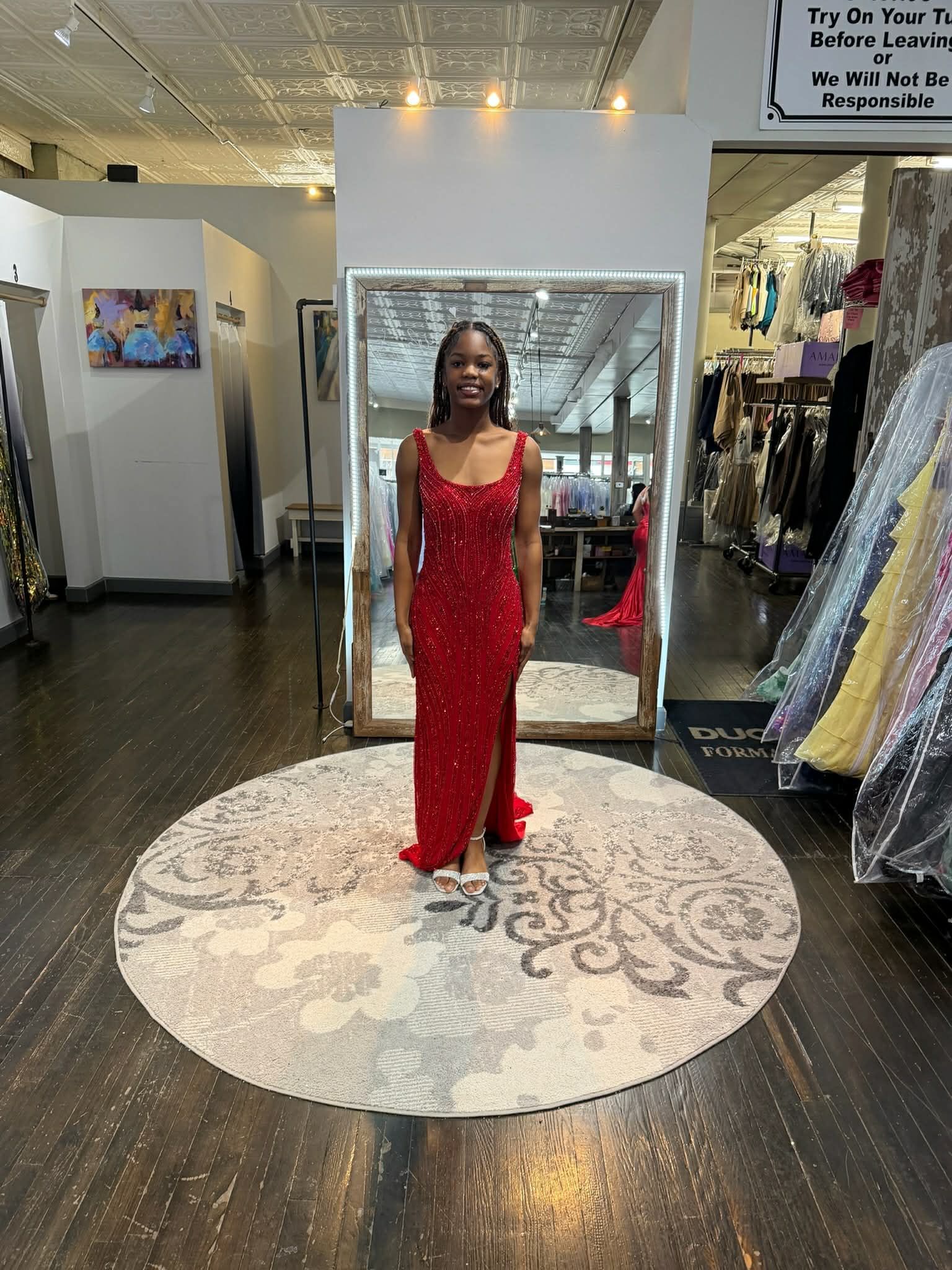 A person stands in a dress shop wearing a floor-length, red, sequined gown with a side slit, positioned on a round rug.