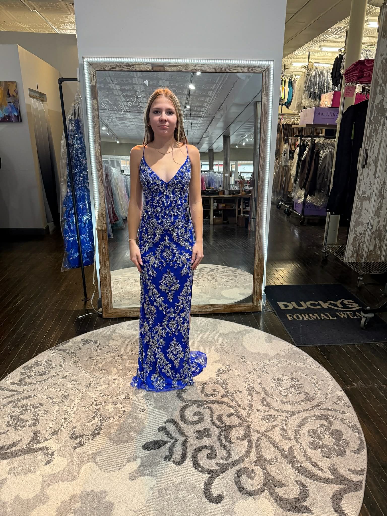 A person stands in a clothing store mirror wearing a floor-length, royal blue beaded gown with a V-neckline.