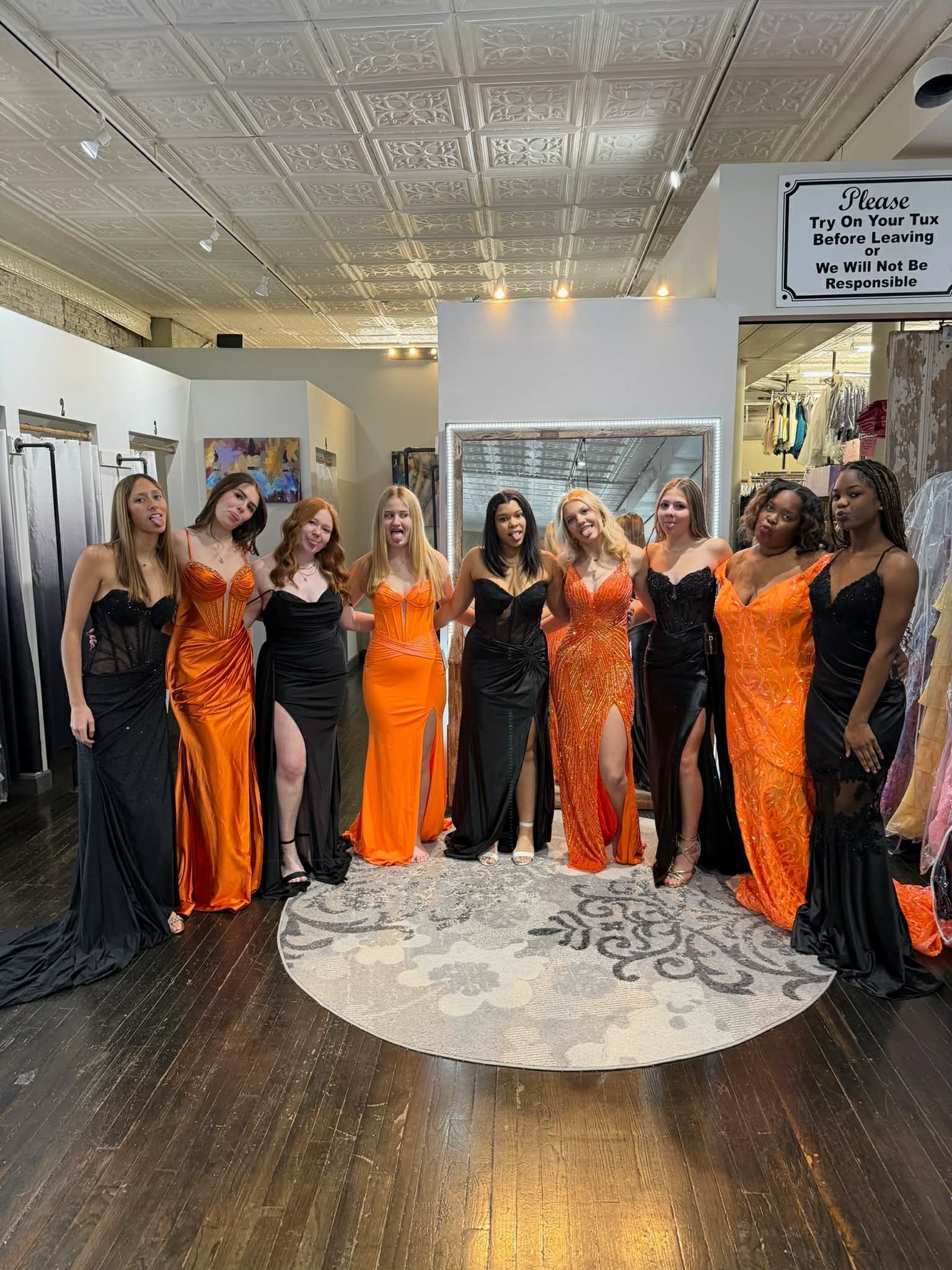 Ten people in long orange and black formal gowns pose together in a clothing boutique with a decorative ceiling.