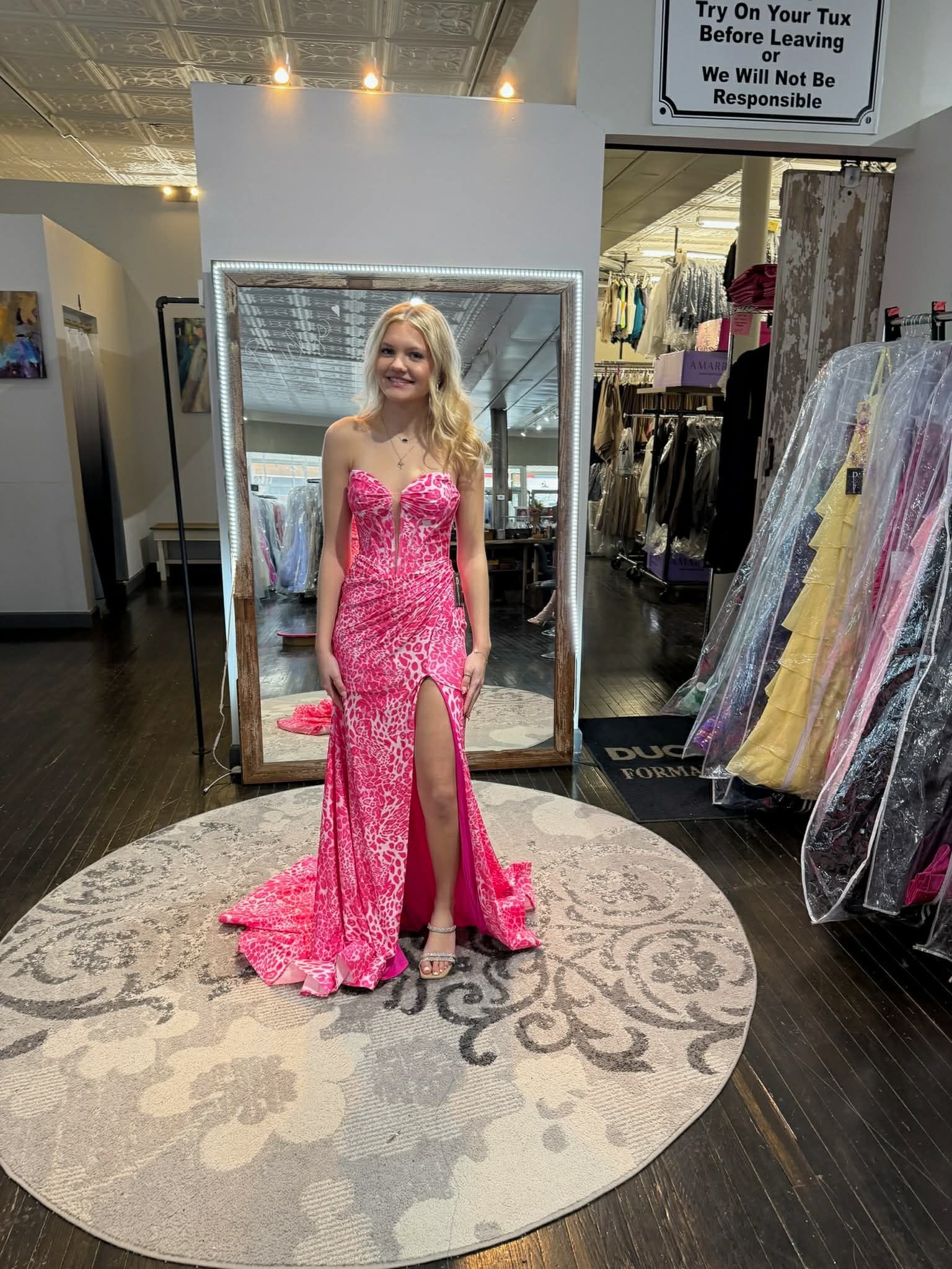 A person stands in a dress shop, smiling while wearing a strapless, pink-and-white patterned gown with a high leg slit.