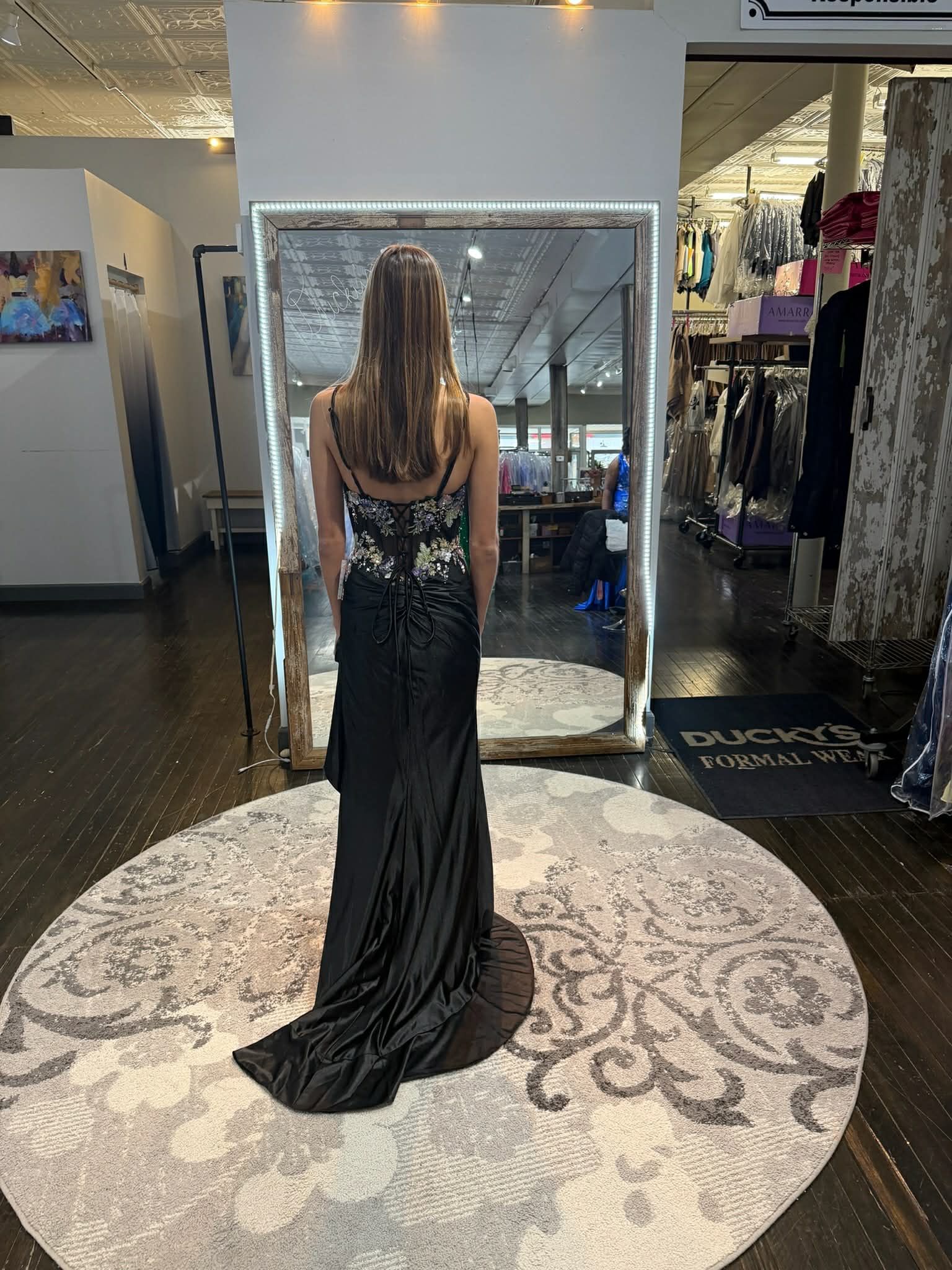 A person wearing a long black evening gown with a sequined bodice stands in a boutique, viewed from behind in a mirror.