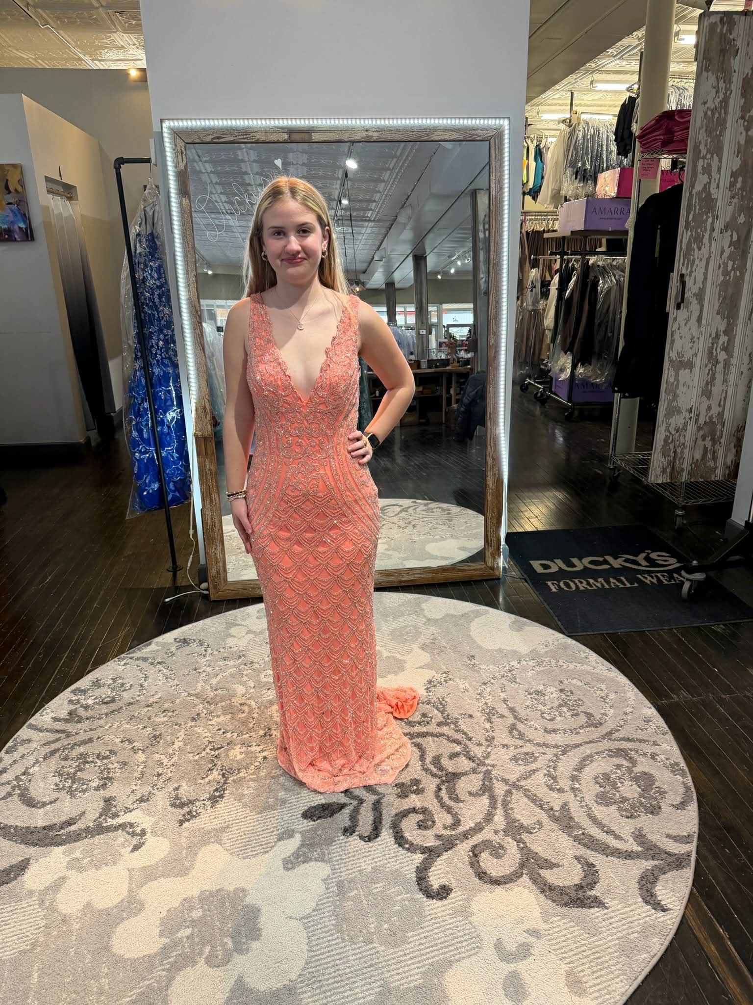 A person standing on a round rug in a clothing store, wearing a floor-length, peach-colored, embellished V-neck gown.