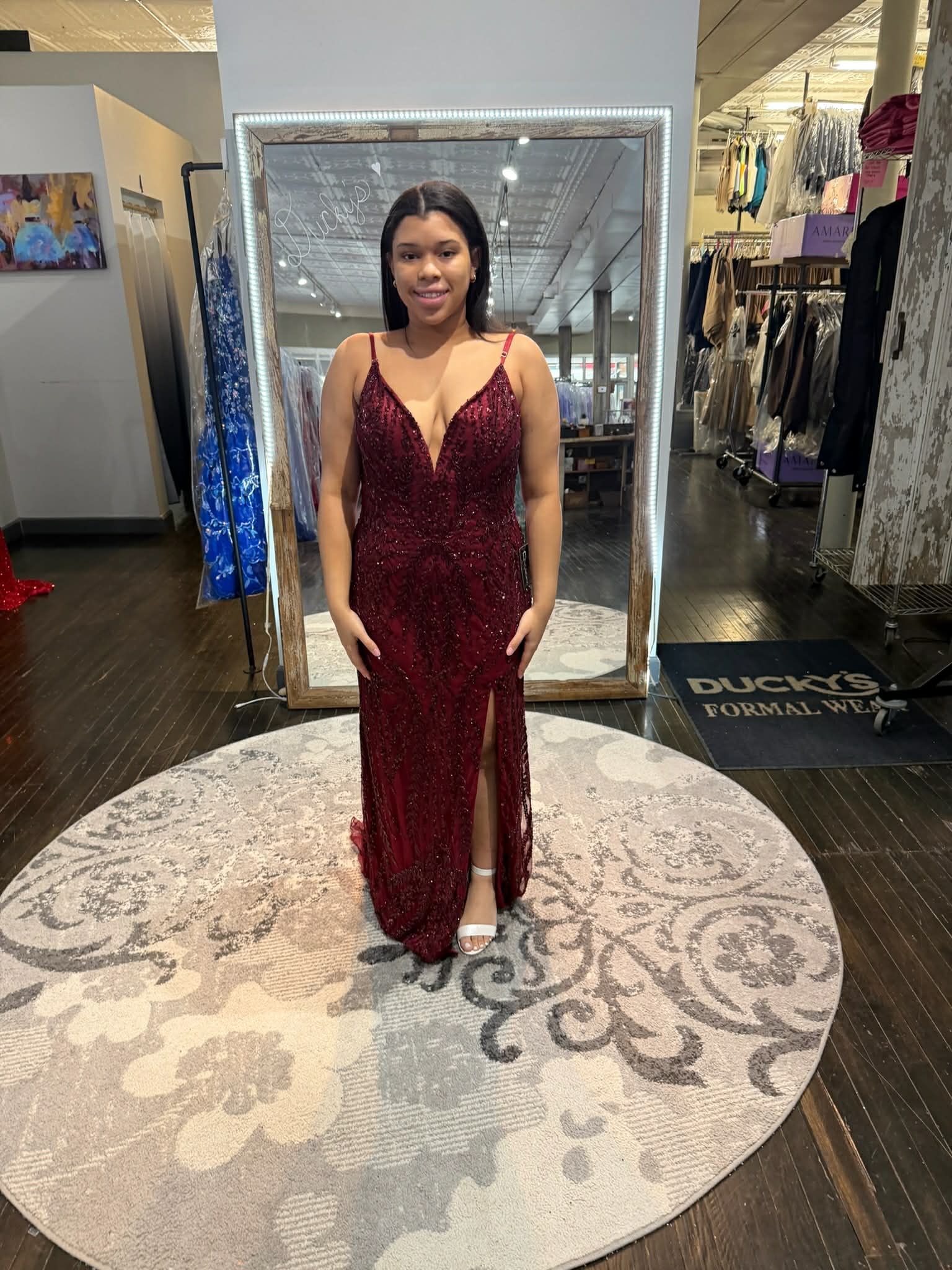 A person stands in a clothing store wearing a floor-length, deep red sequined gown with a slit, centered in a mirror.