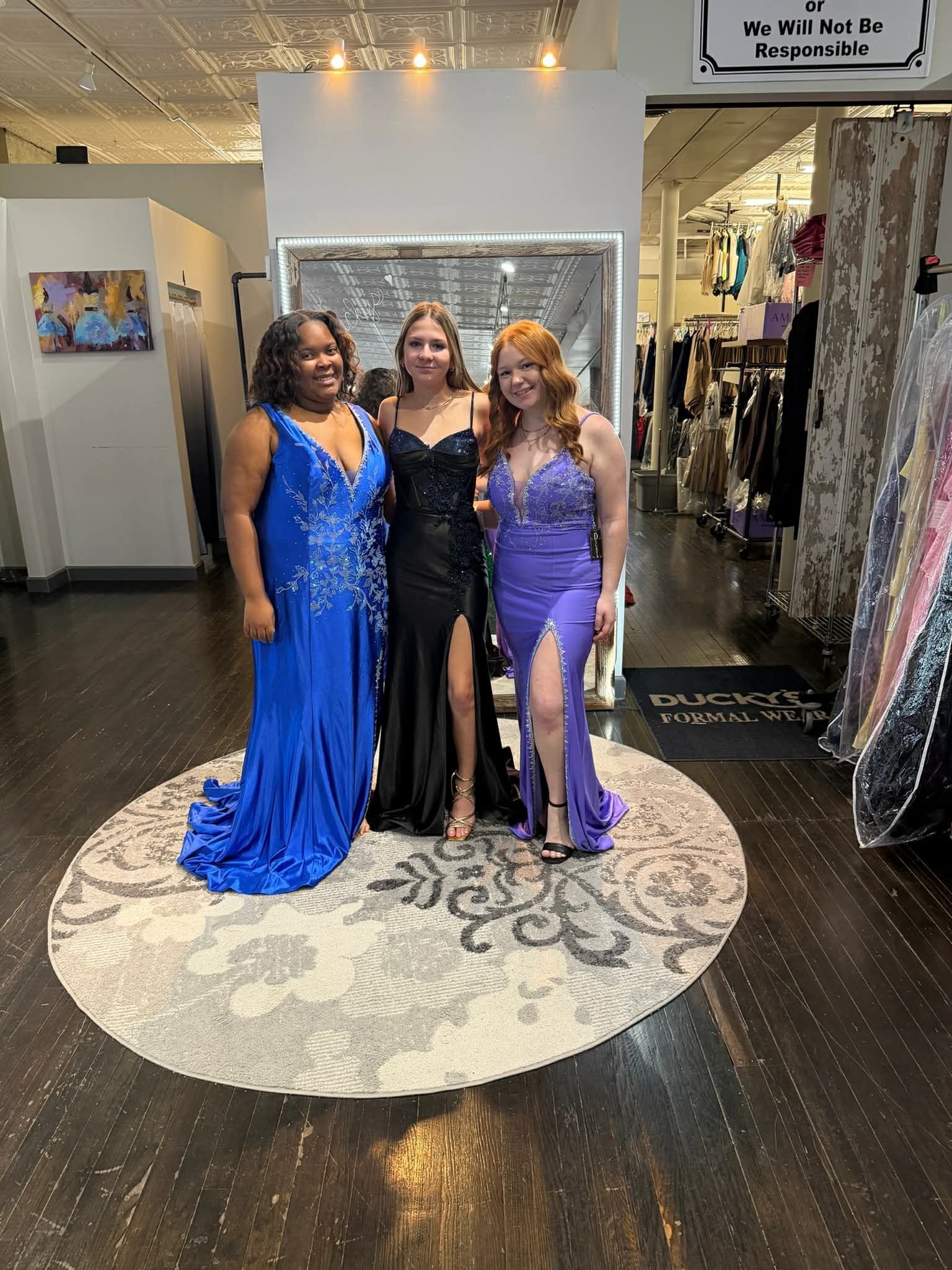 Three people stand in a clothing store, posing in formal gowns of blue, black, and light purple on a circular rug.