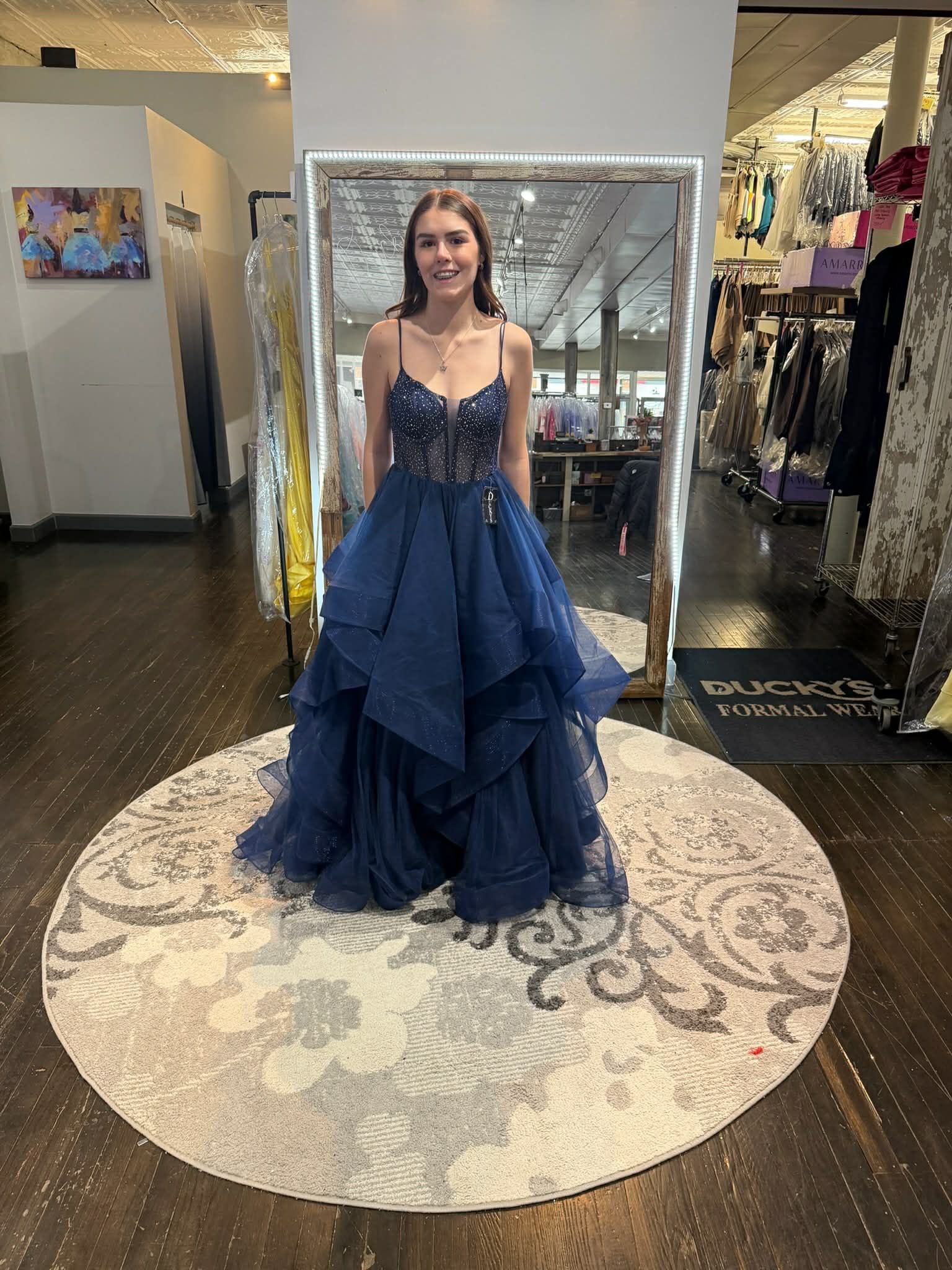 A person smiles while wearing a navy blue, tiered tulle ballgown in a boutique.