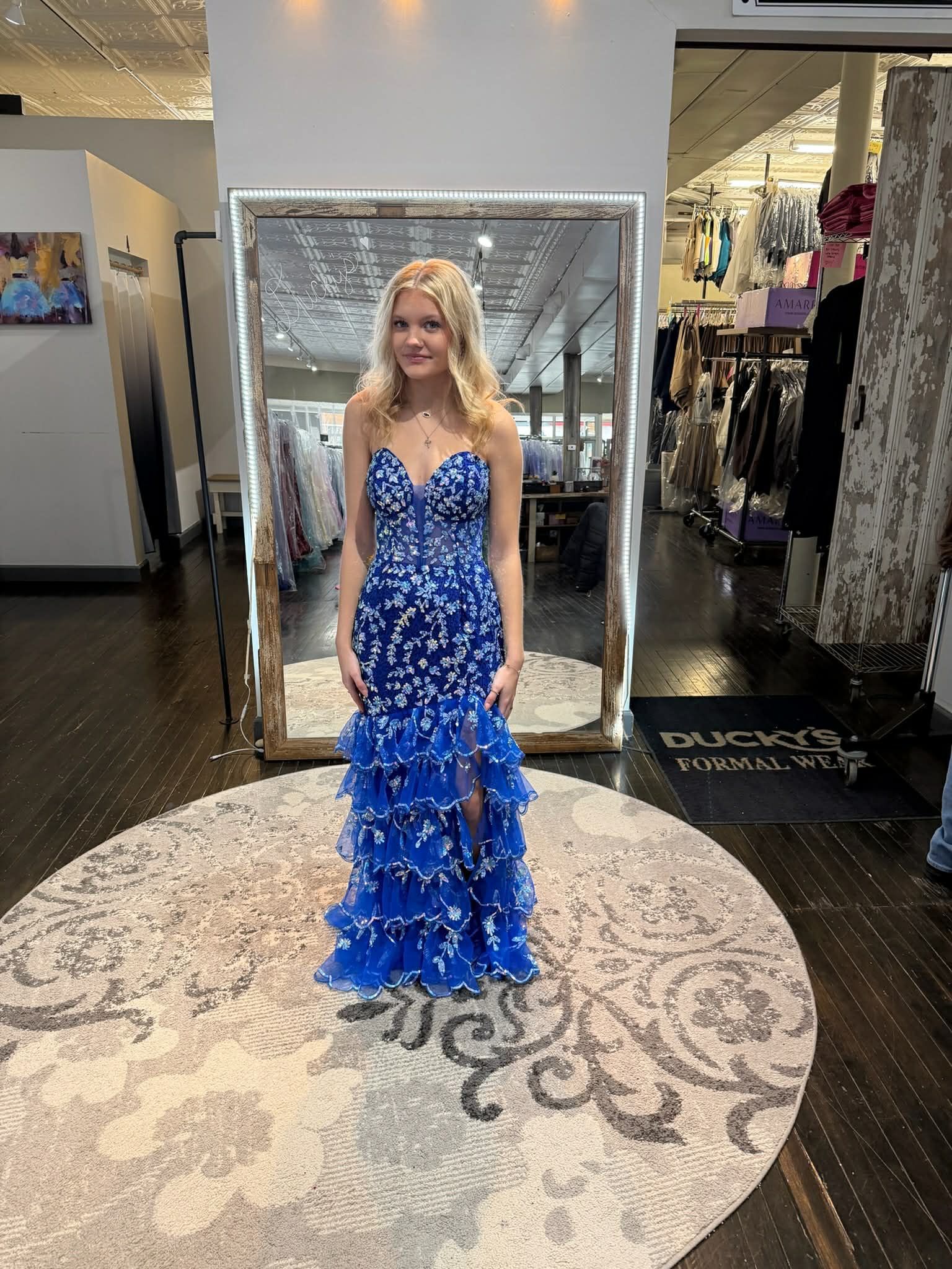 A person stands in a clothing boutique wearing a royal blue, floor-length gown with a sequined bodice and tiered skirt.