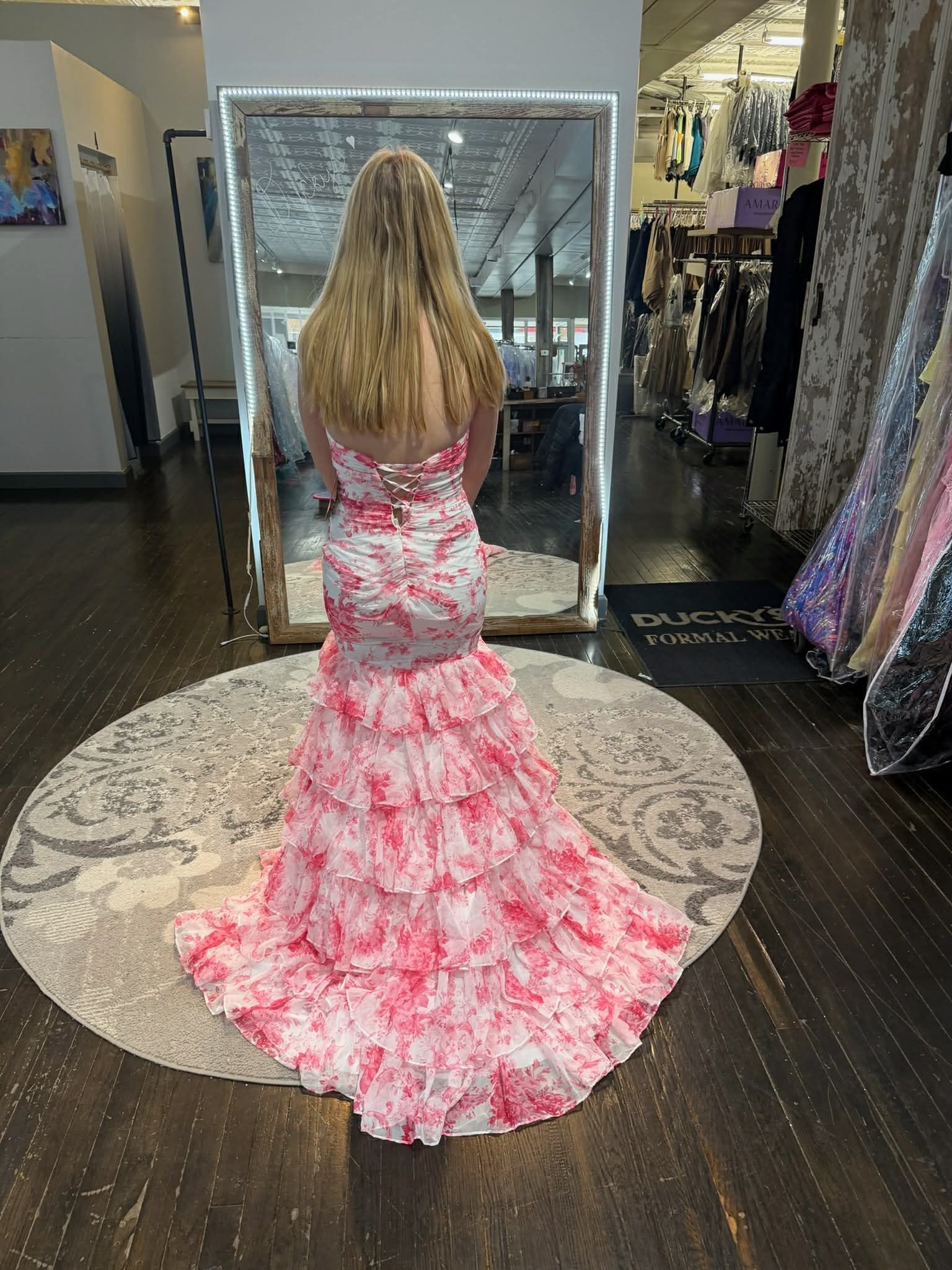 A person stands in a dress shop, seen from behind, wearing a white, tiered, floral-patterned gown in shades of pink.