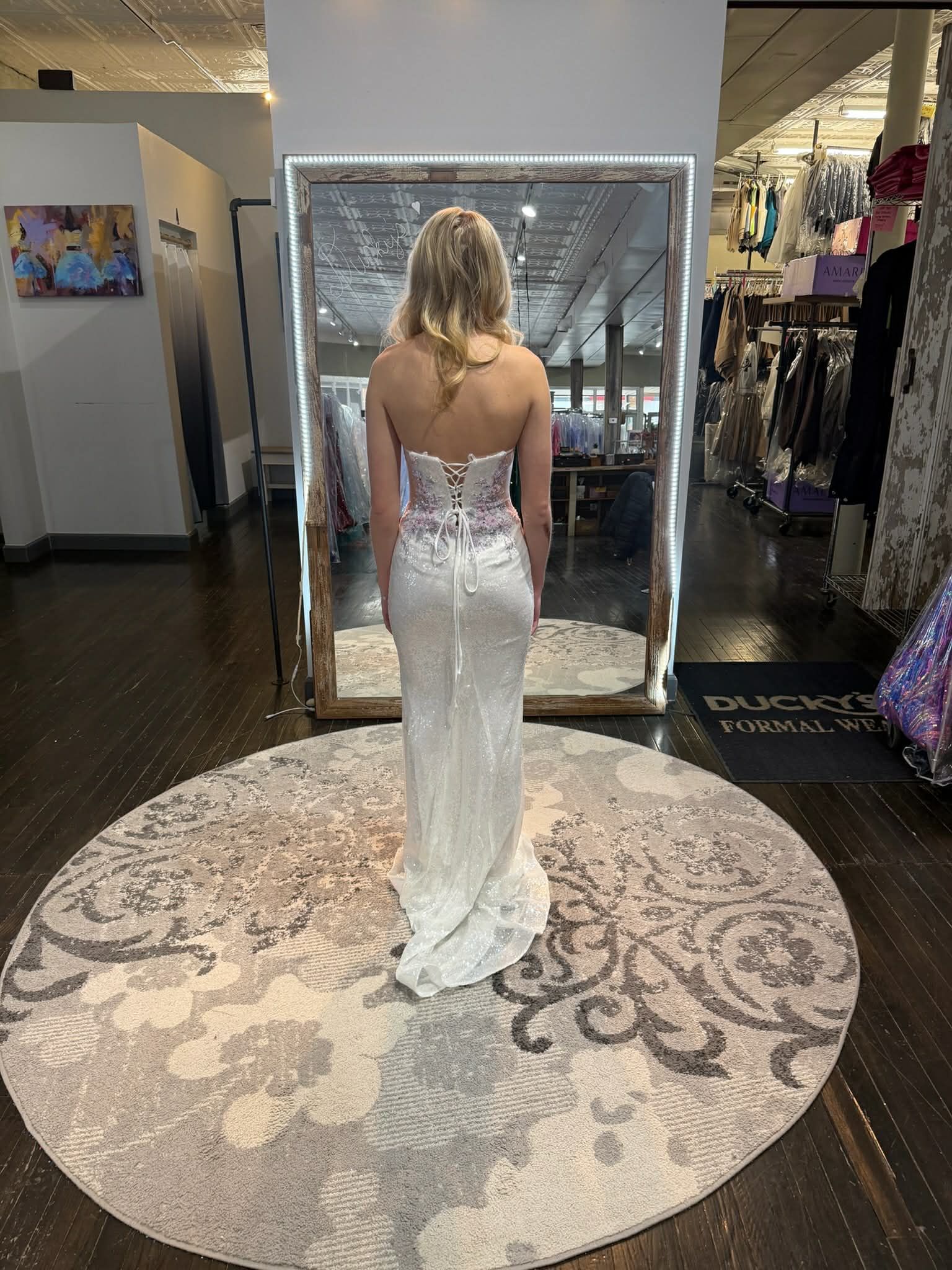 A person wearing a white, sequined, lace-up back floor-length gown stands in front of a mirror in a clothing store.