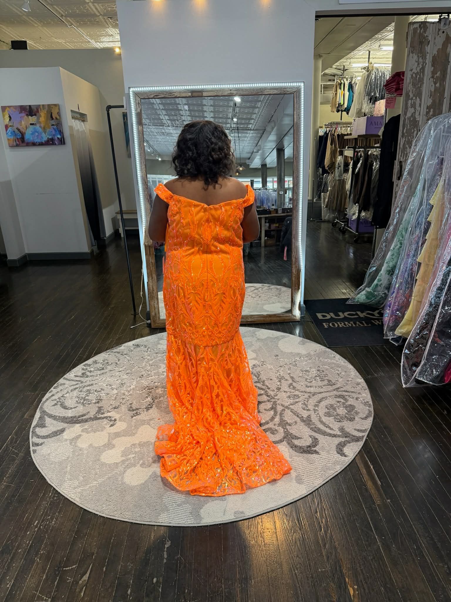 A person stands in a dress shop wearing a bright orange, sequined off-the-shoulder mermaid-style gown.