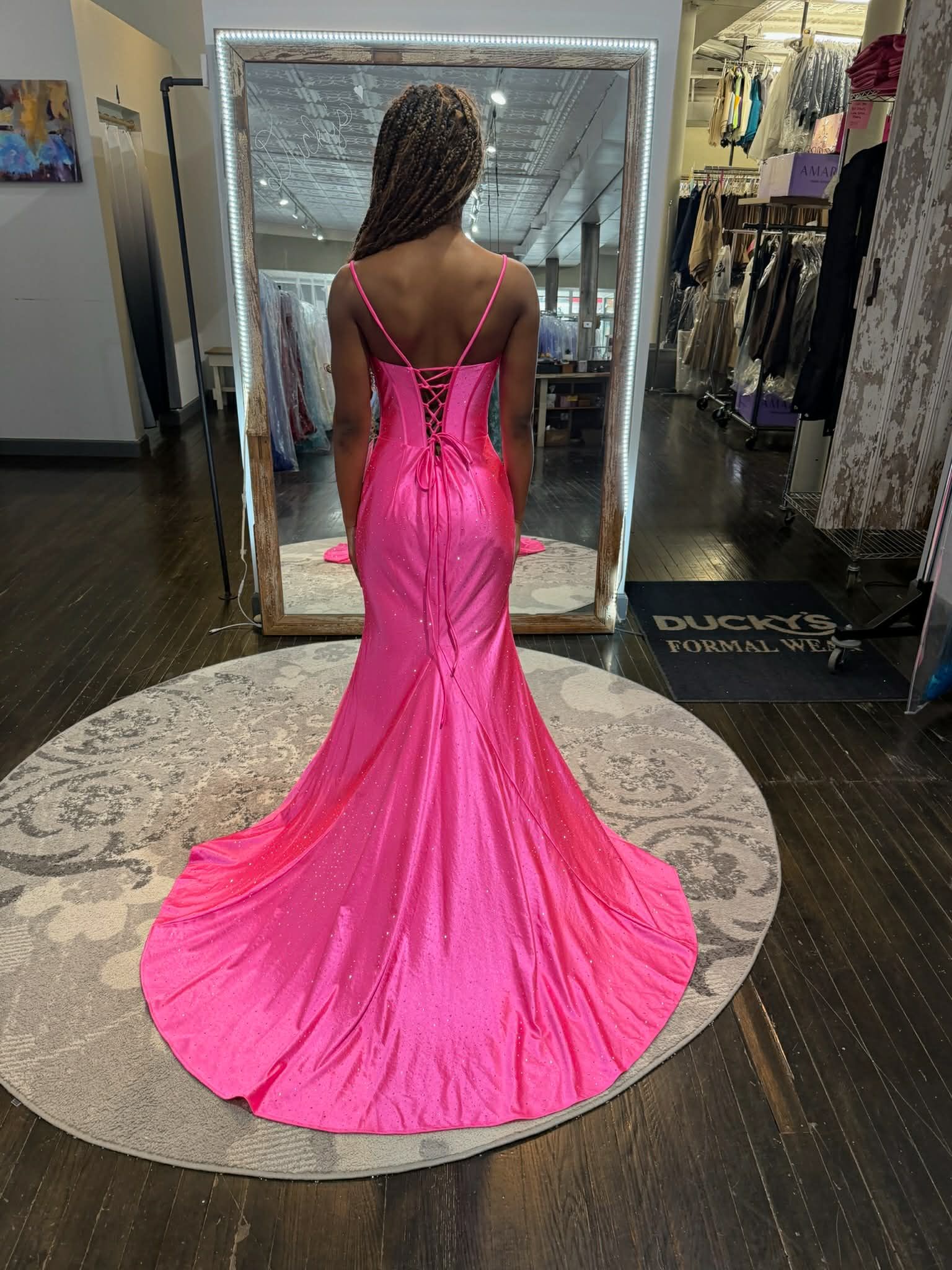 A person in a bright pink, floor-length gown with a lace-up back and train, standing on a rug in a dress shop.