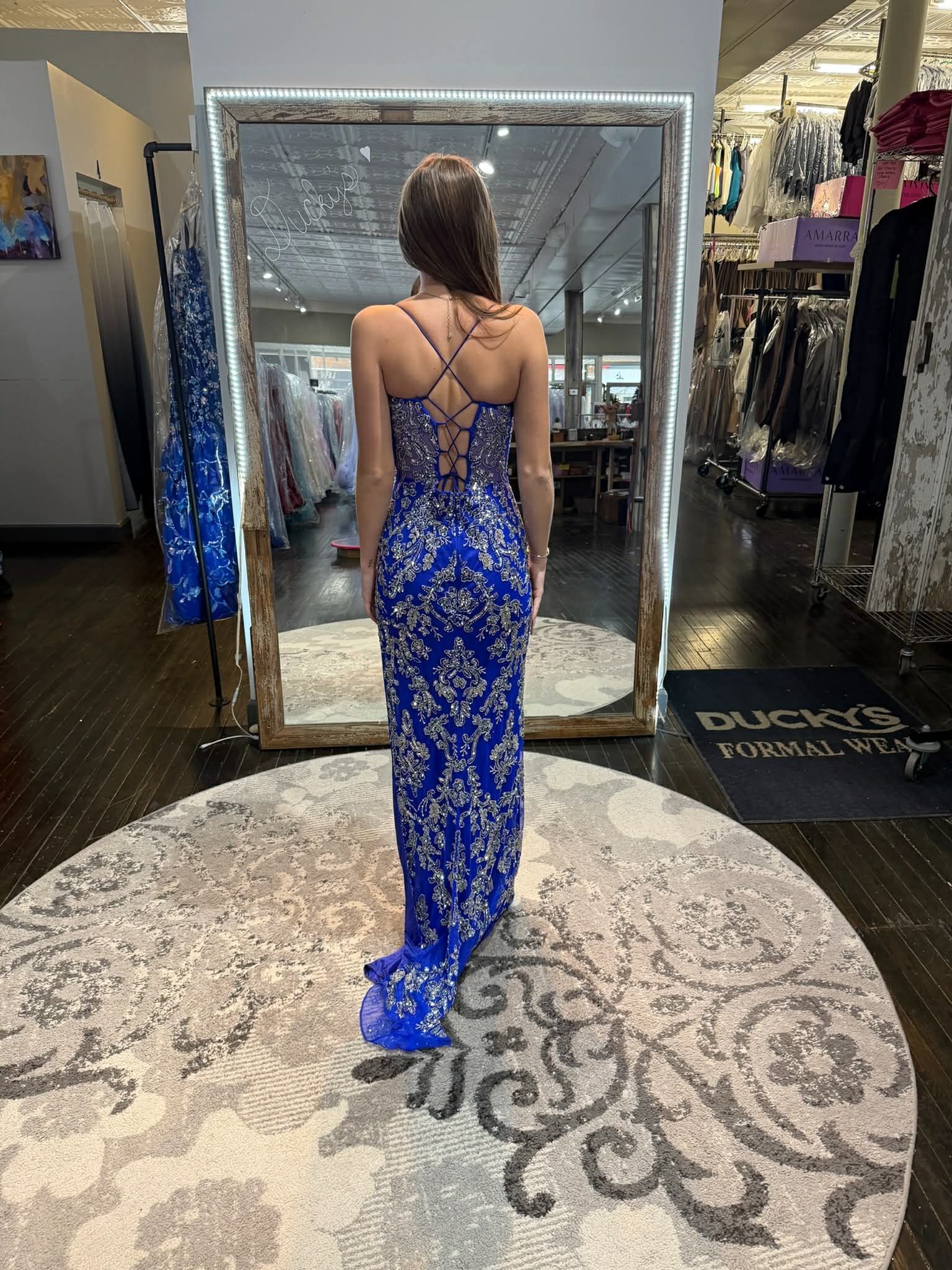 A person stands in a clothing store, facing a mirror while wearing a floor-length, blue dress with silver floral detailing.