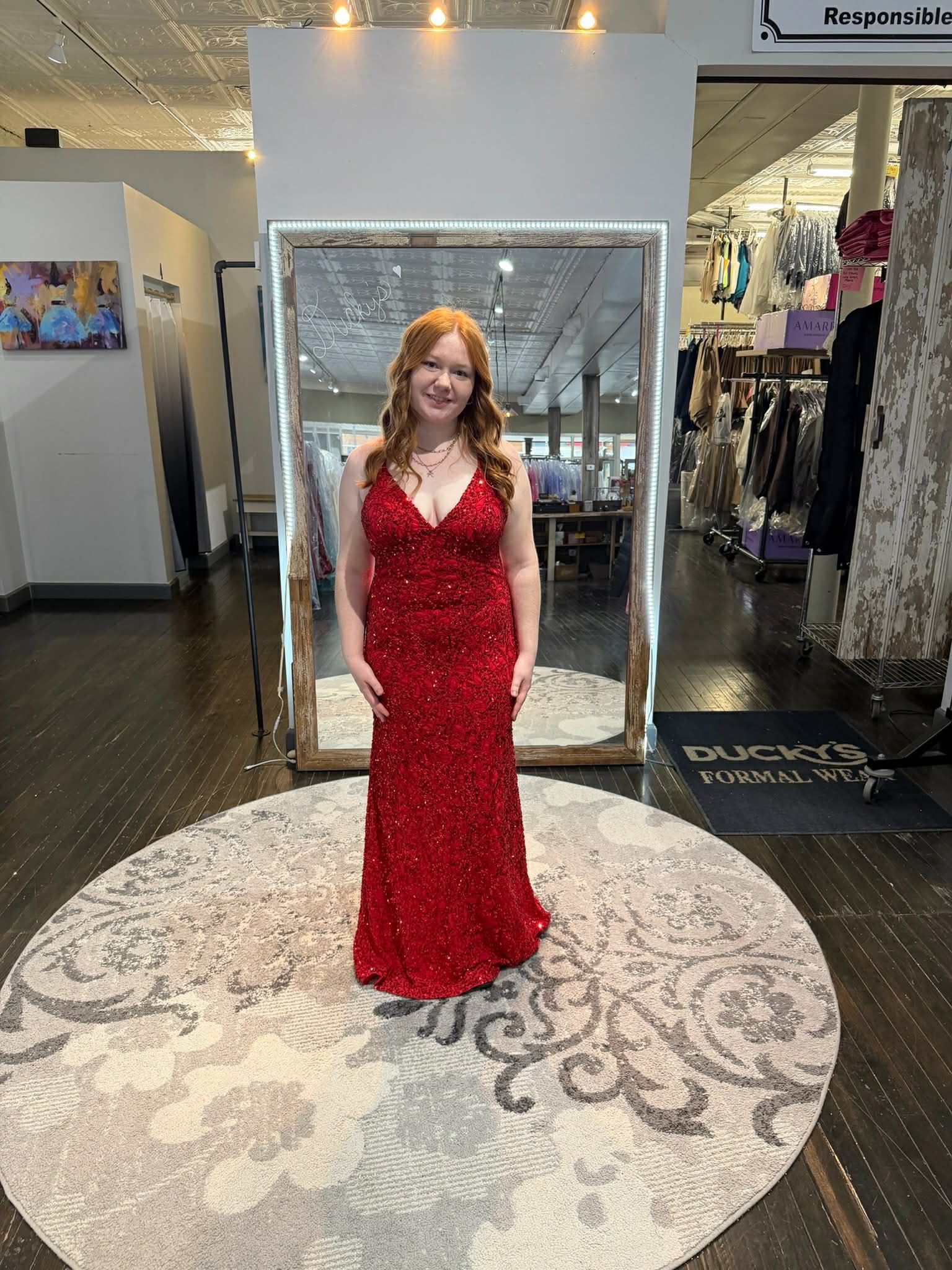 A person smiles in a full-length, sparkling red gown, standing on a circular rug in a formal wear clothing store.