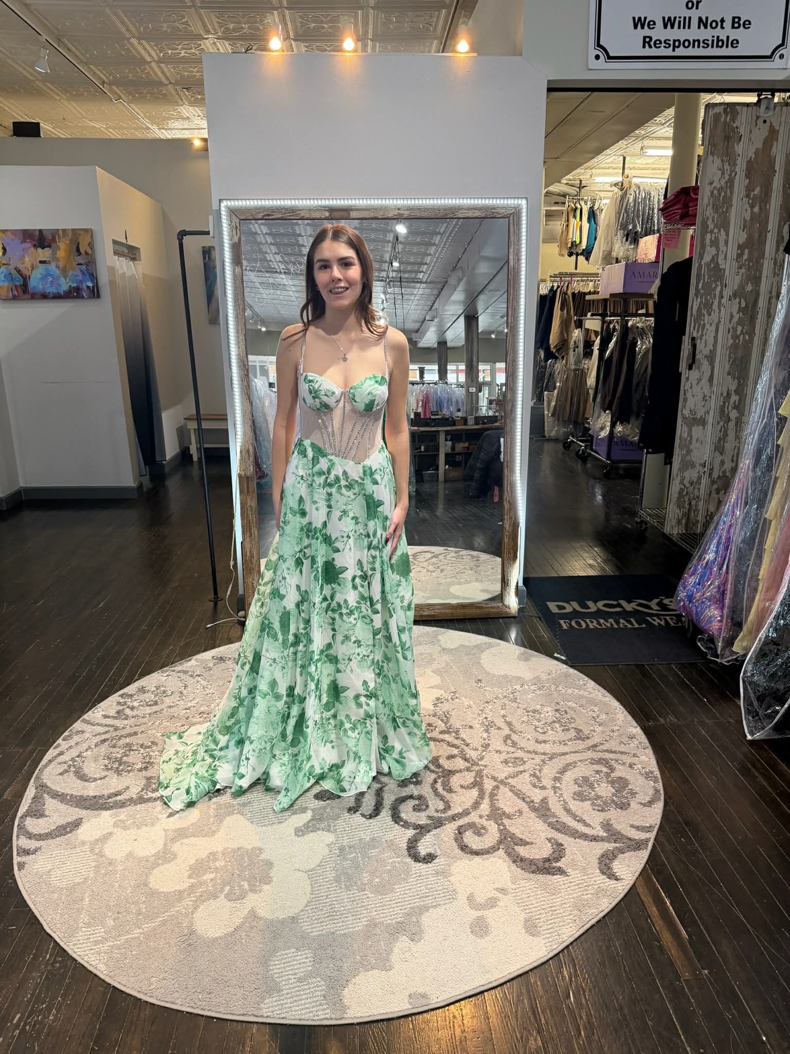 A person wearing a floral green and white gown with a lace corset bodice stands in a boutique store in front of a mirror.