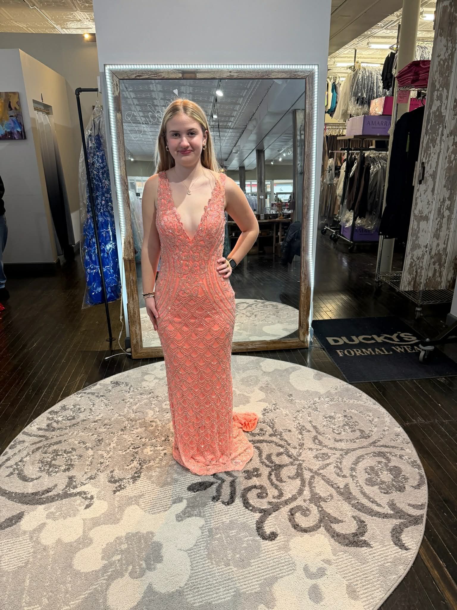 A person wearing a sparkling peach-colored, floor-length gown stands in a clothing boutique in front of a floor mirror.