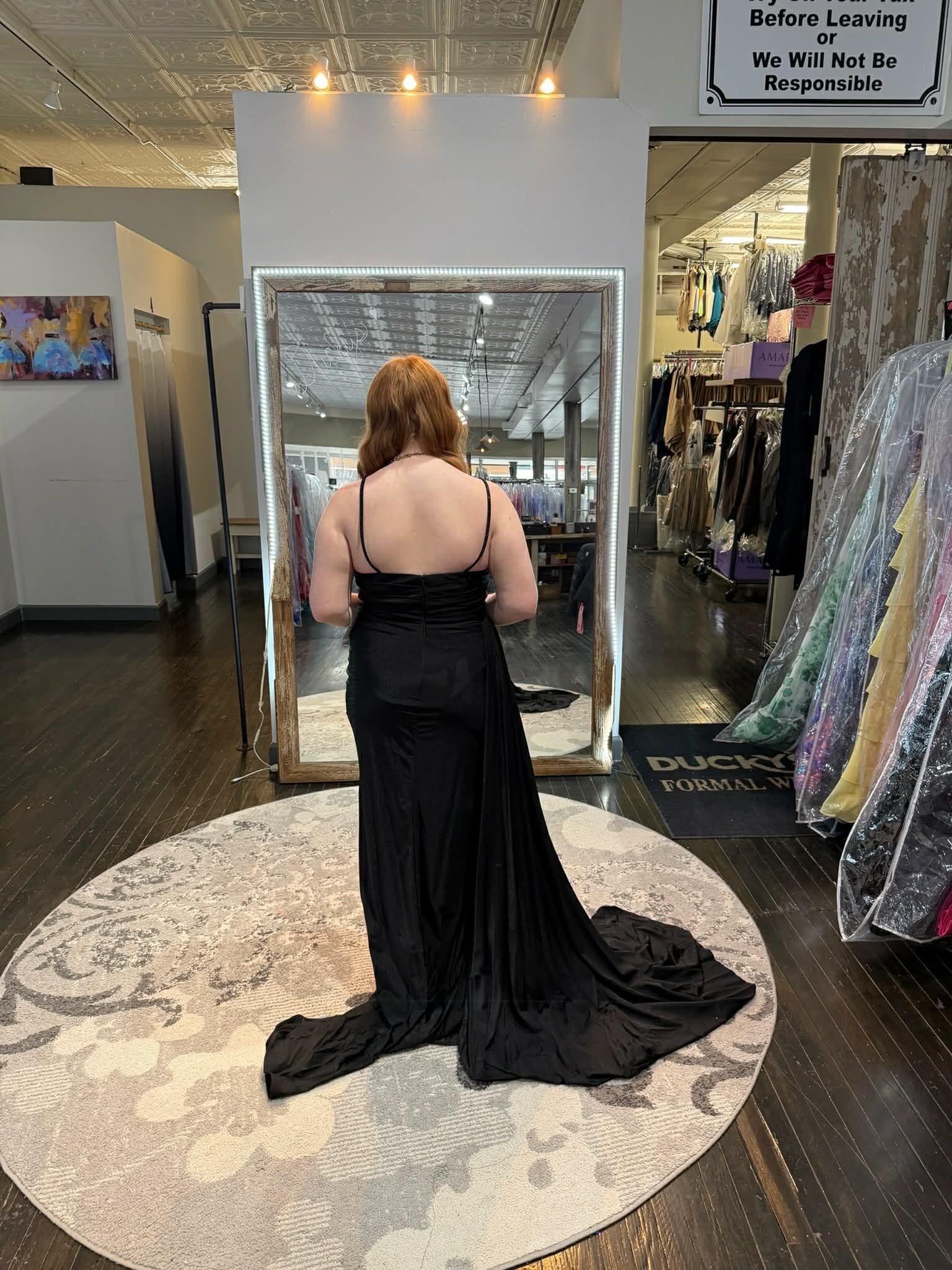 A person wearing a long black dress stands on a circular rug in front of a mirror in a clothing store.