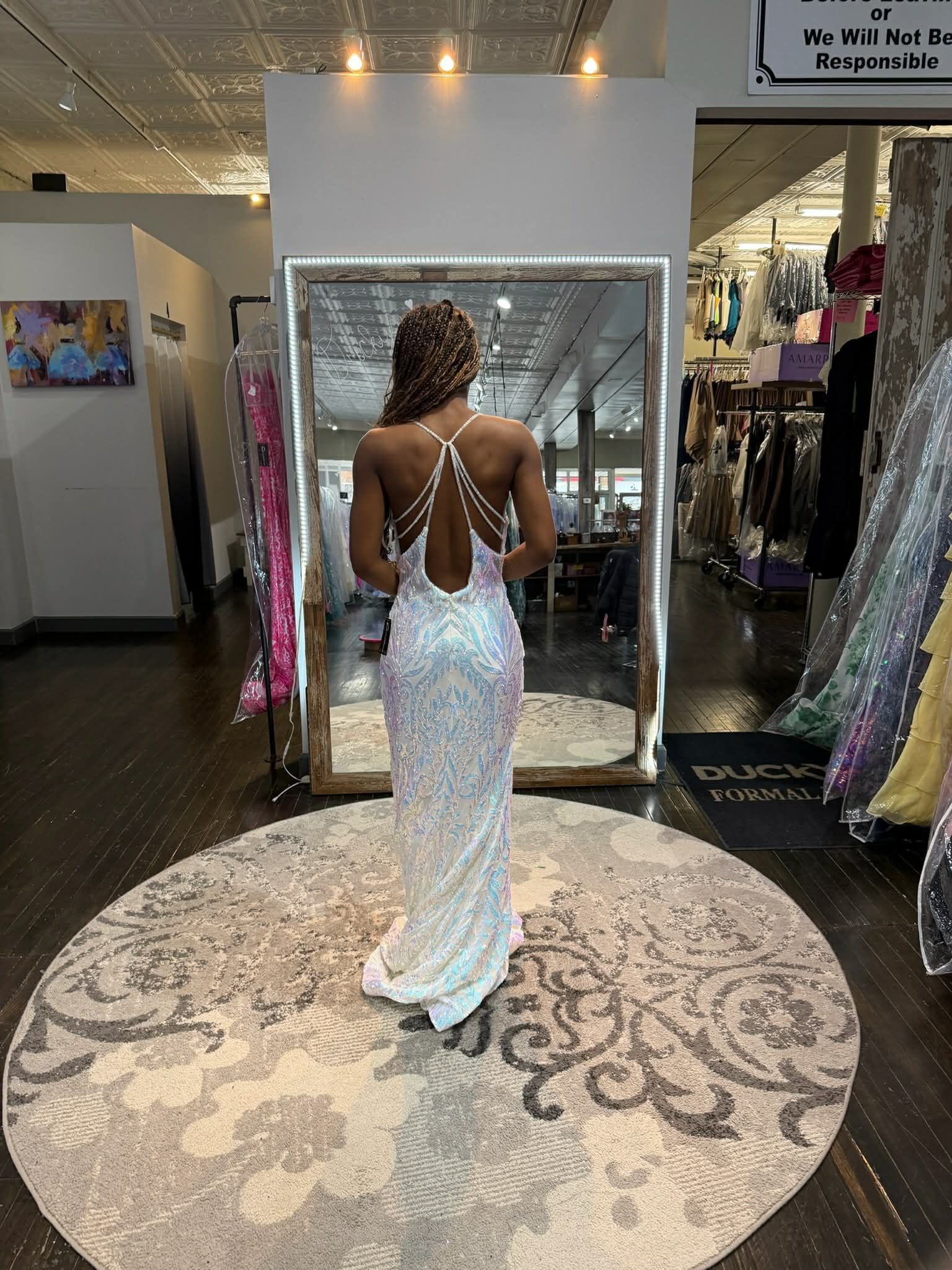 A person wearing a floor-length, backless white sequined gown stands on a round patterned rug in a clothing store.