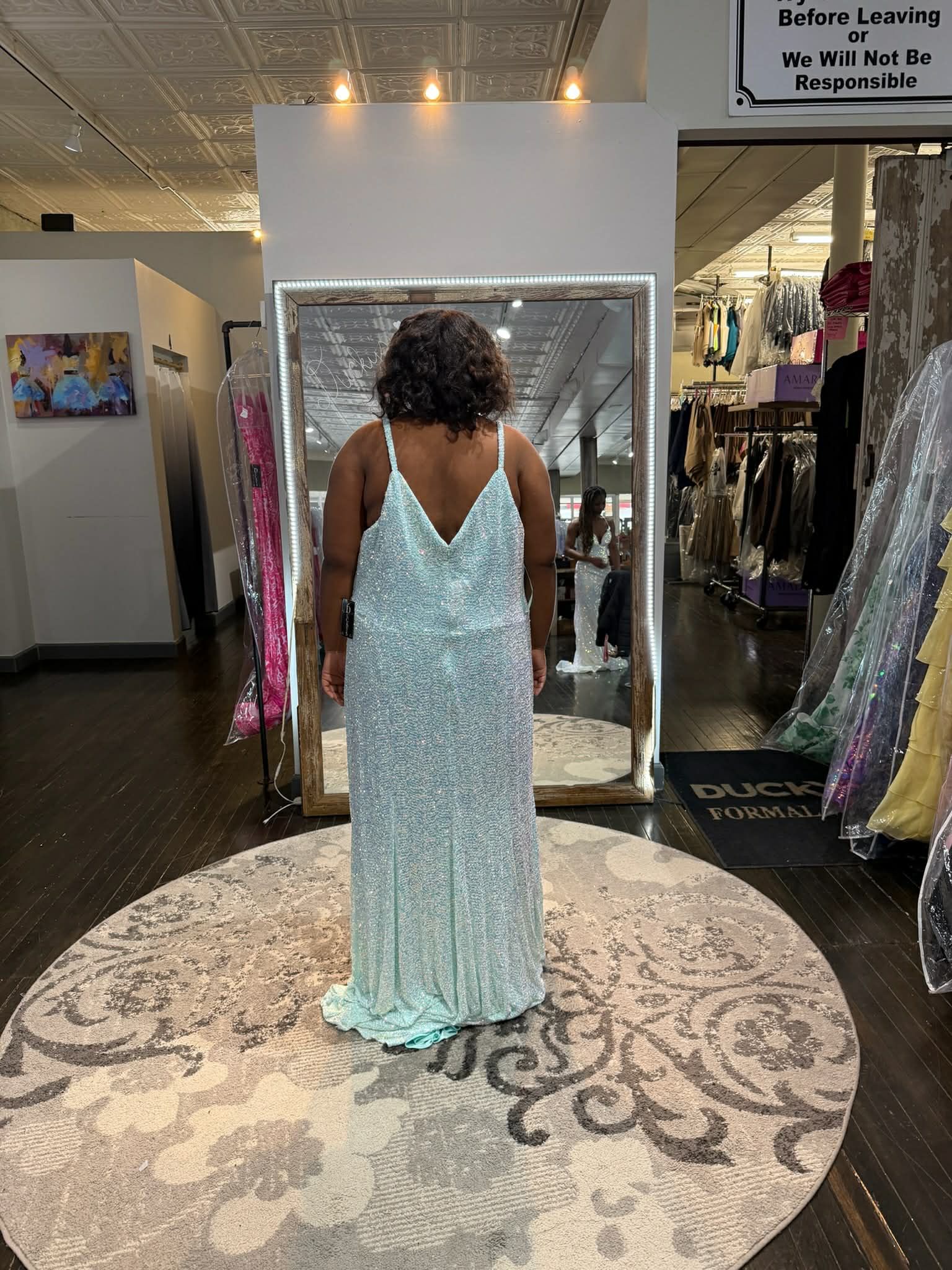 A person wears a sparkly, light blue floor-length gown with thin straps while standing on a circular rug in a boutique.