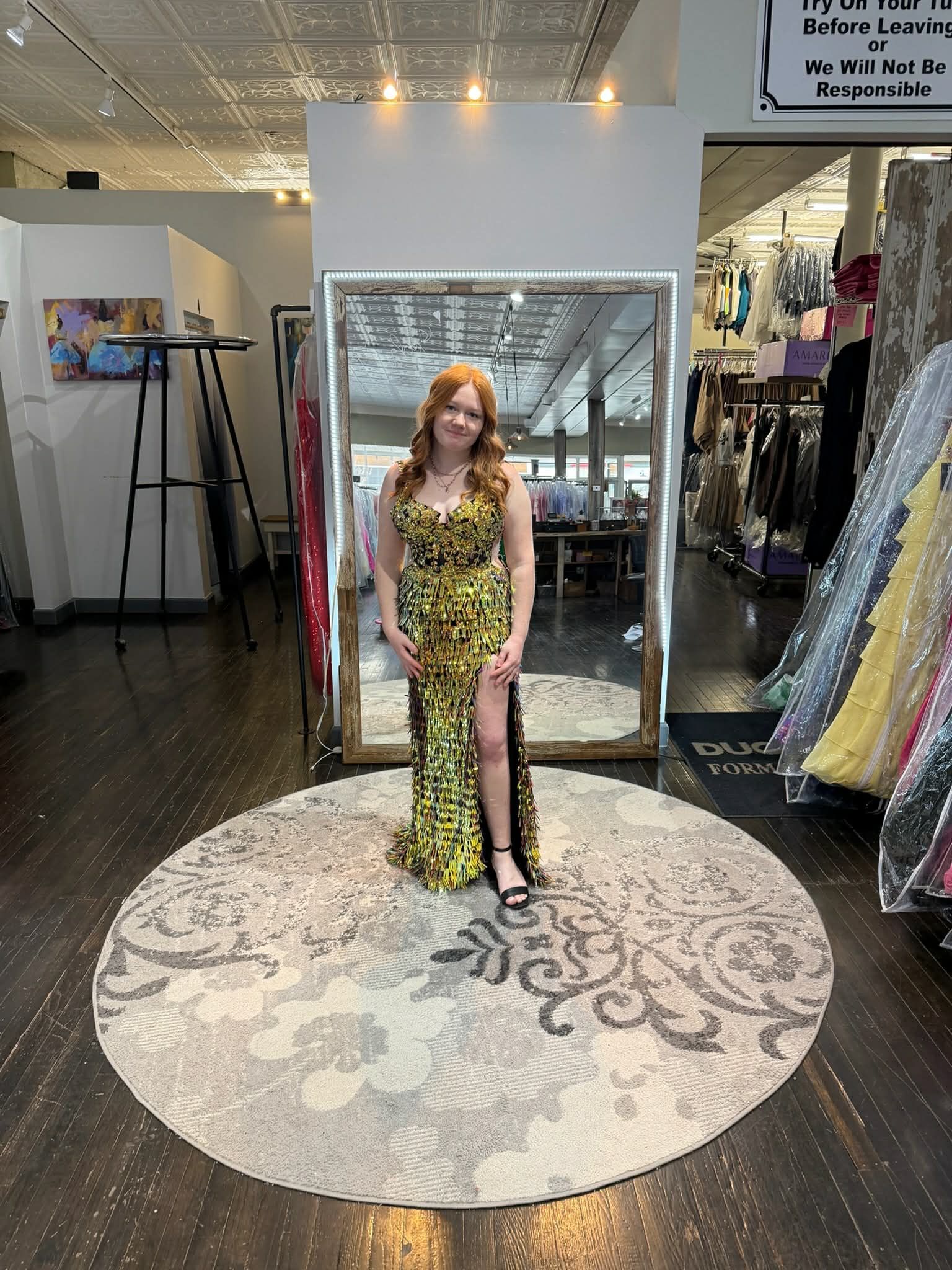 A person wearing a shimmering gold sequin floor-length dress with a side slit stands on a round rug in a clothing store.