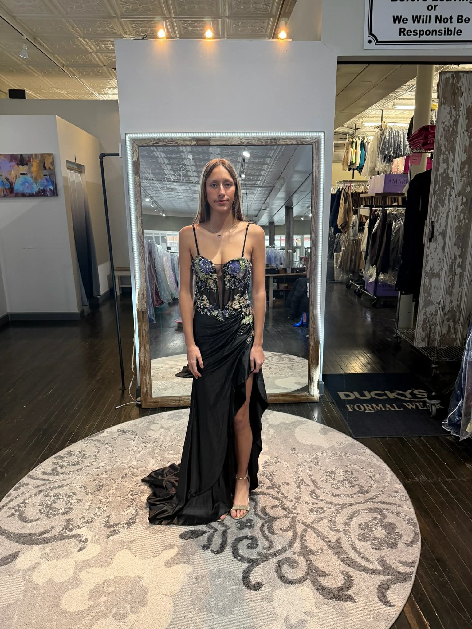 A person stands in a clothing store mirror wearing a floral corset top with a black, high-slit skirt and a train.