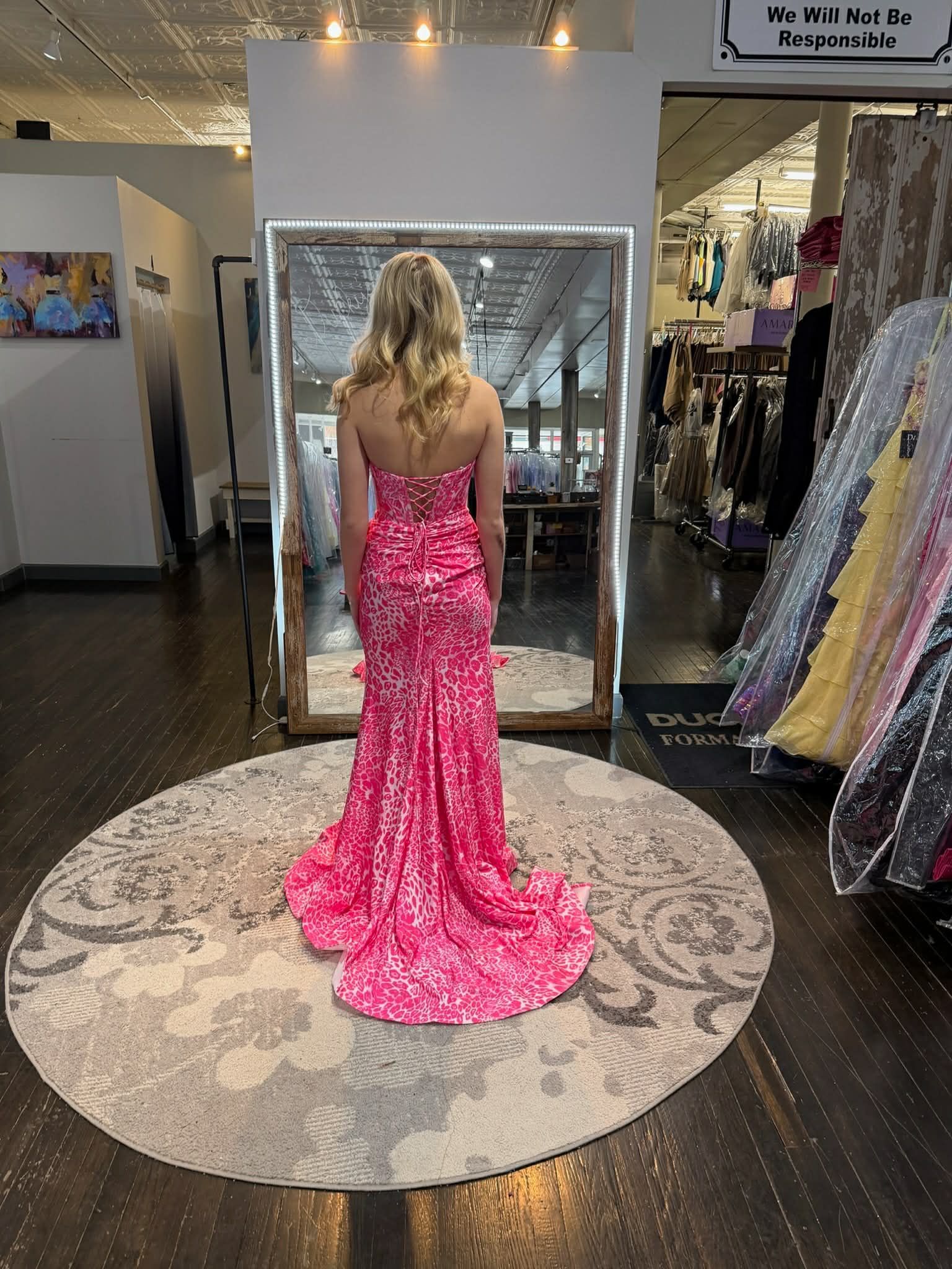 A person wearing a strapless, pink patterned floor-length gown with a small train, standing on a circular rug in a store.
