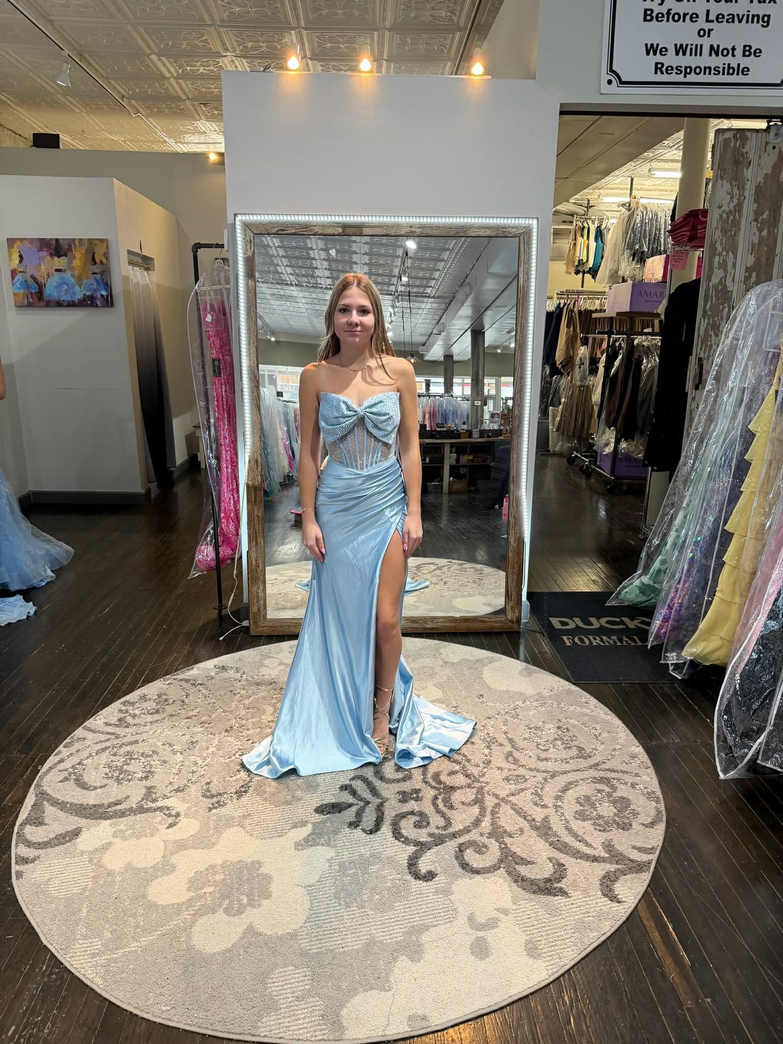 A person stands in a dress shop, modeling a light blue, floor-length gown with a side slit and floral bodice detail.