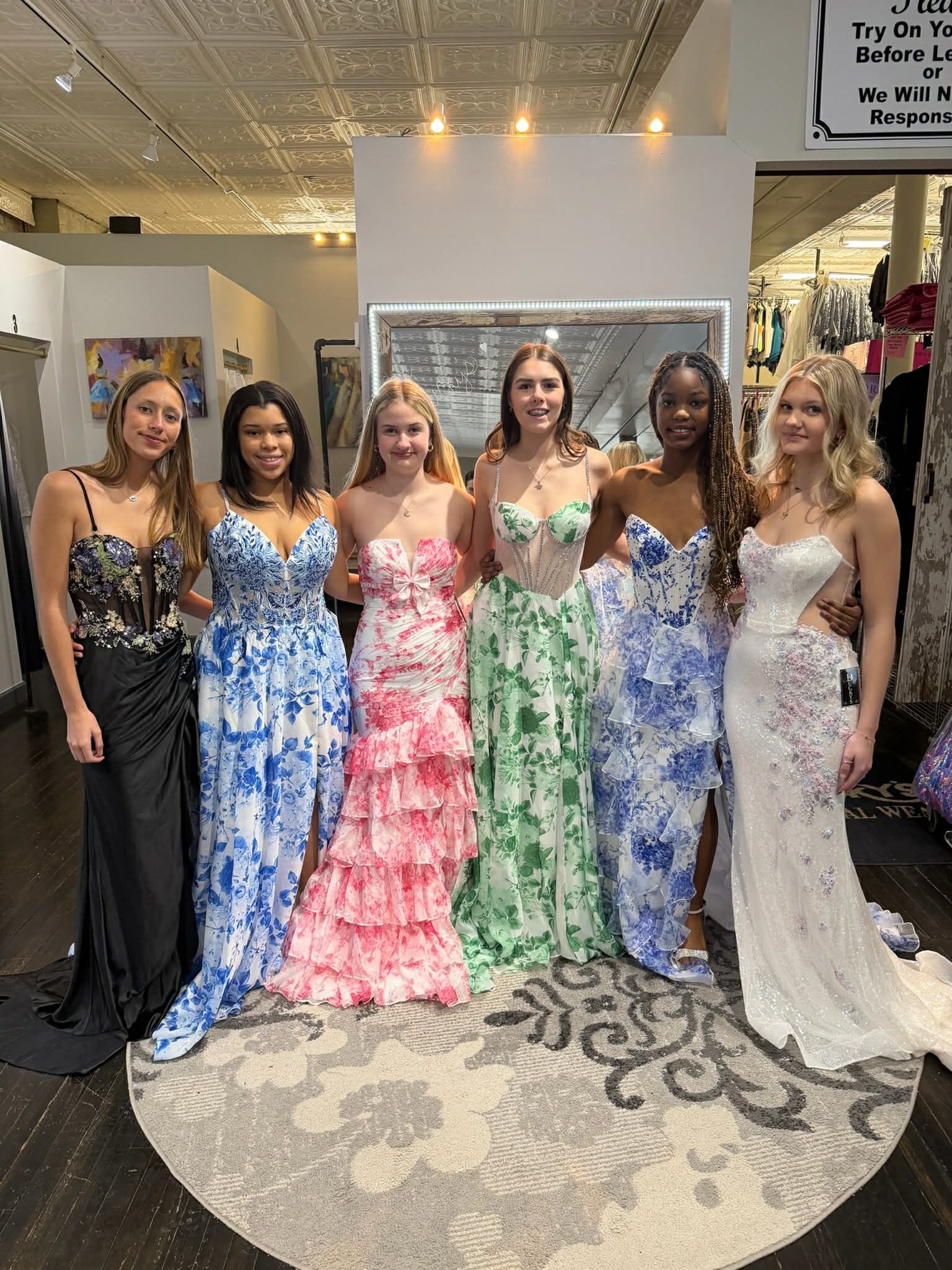Six people stand in a boutique, posing in various colorful and floral-patterned formal gowns on a circular rug.