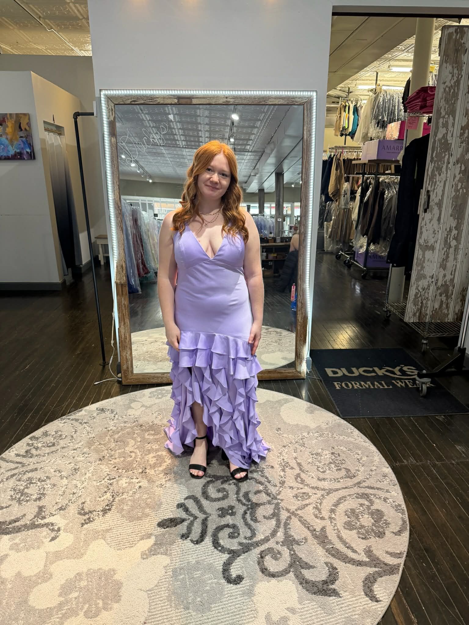 A person with red hair smiles while wearing a floor-length lavender ruffled gown in a clothing store with a large mirror.