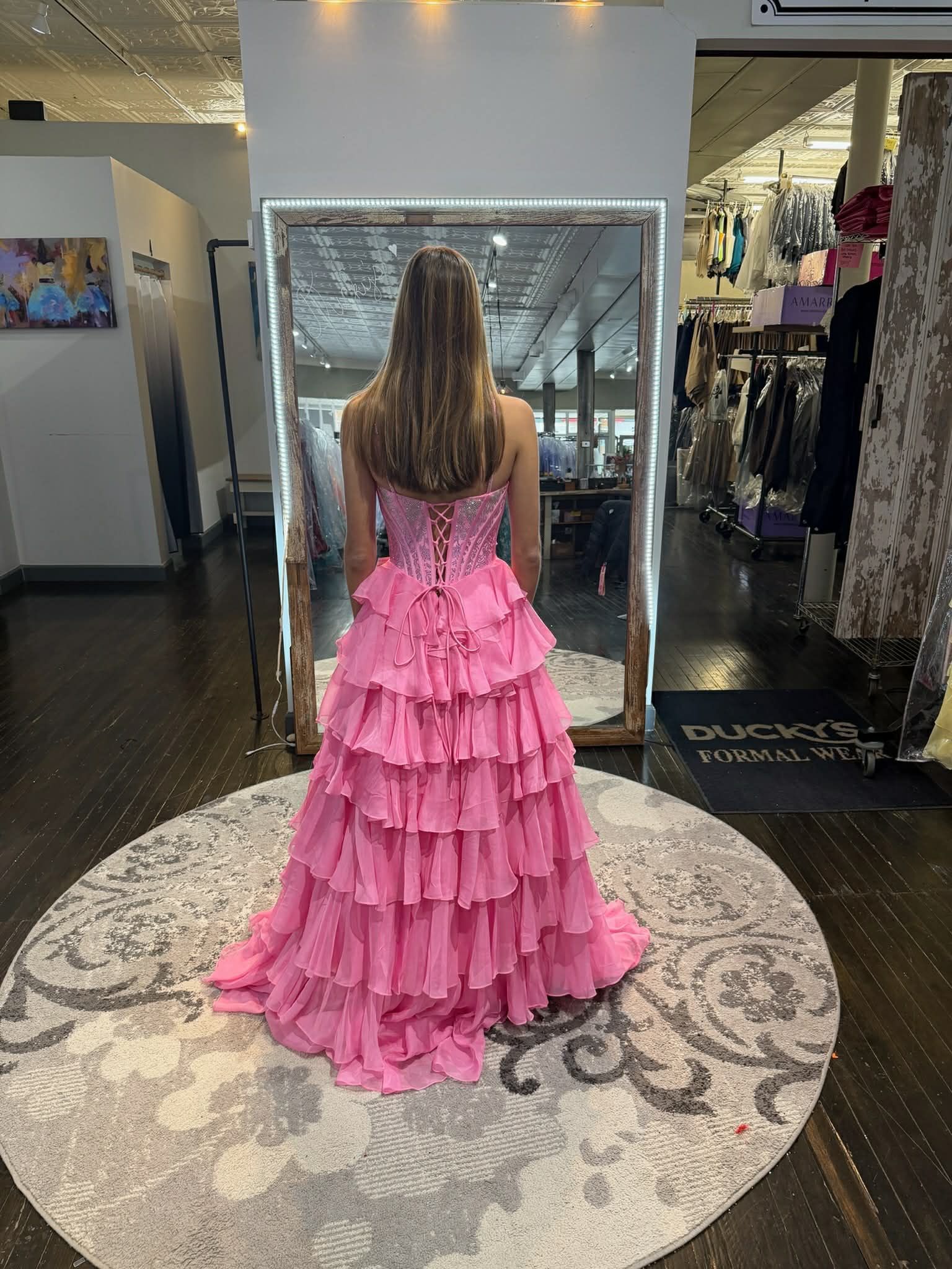 A person stands in a clothing store mirror wearing a bright pink, floor-length gown with a tiered ruffle skirt.