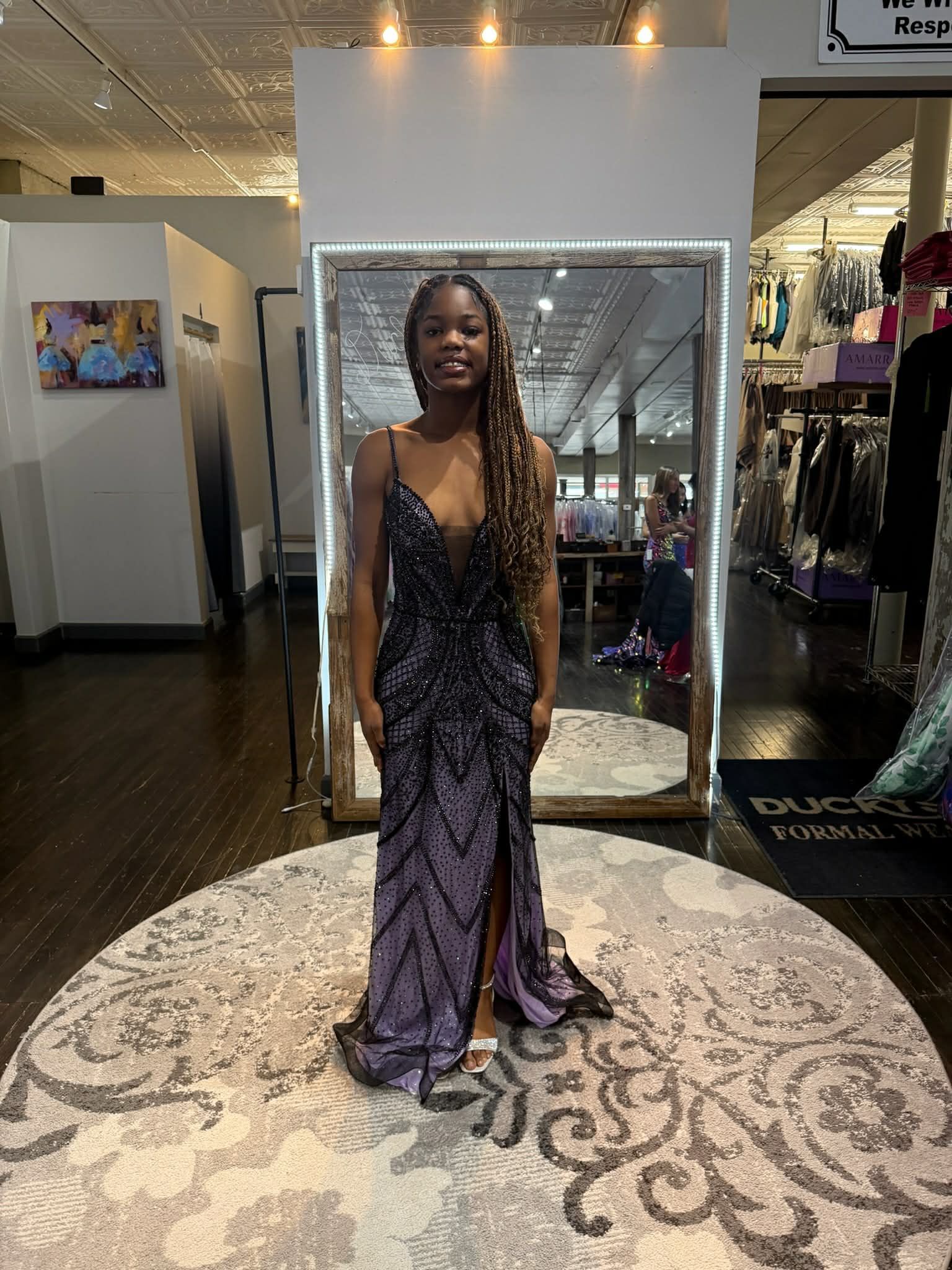A person stands in a clothing boutique wearing a floor-length, purple, sequined gown with a high slit.