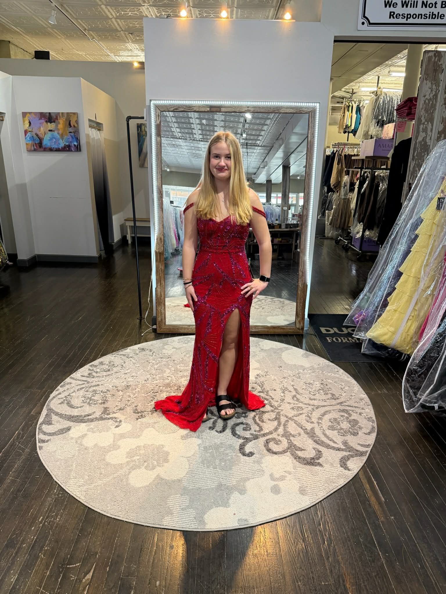 A person wearing a long, red sequined evening gown with a high slit stands on a round patterned rug in a clothing store.