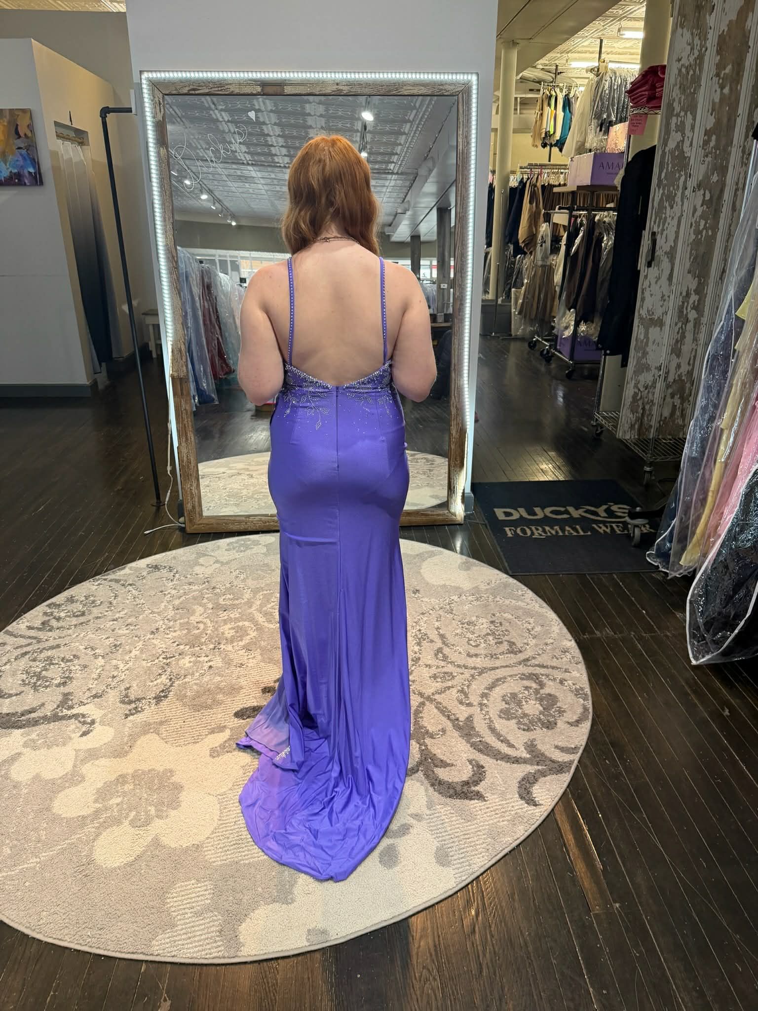 A person standing in front of a mirror wearing a floor-length, backless purple gown with embellished straps in a shop.