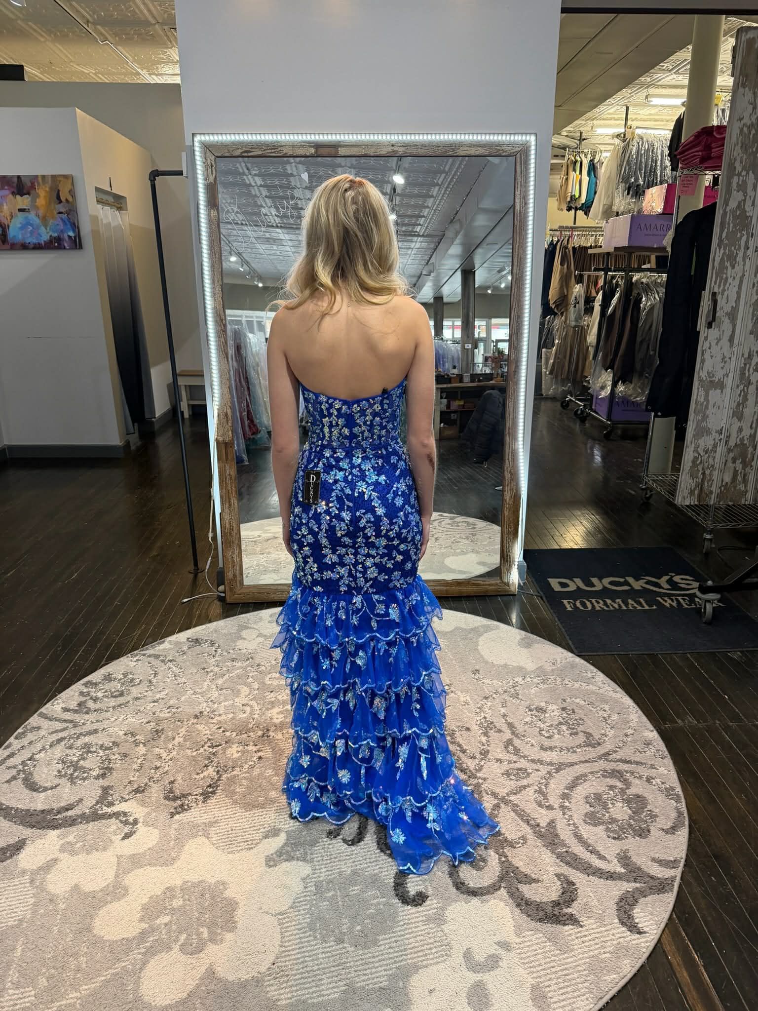 A person viewed from behind, standing on a rug in a dress shop while wearing a blue, strapless, tiered evening gown.