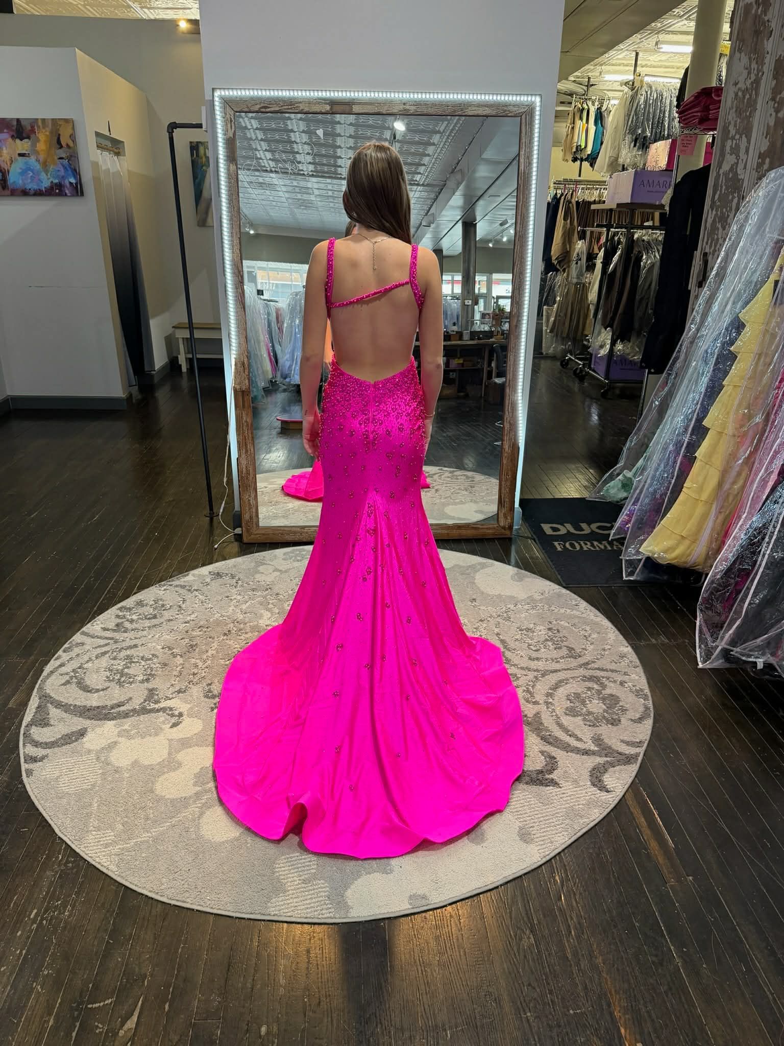 A person stands in a dress shop wearing a bright pink, floor-length sequined gown with an open back and a train.