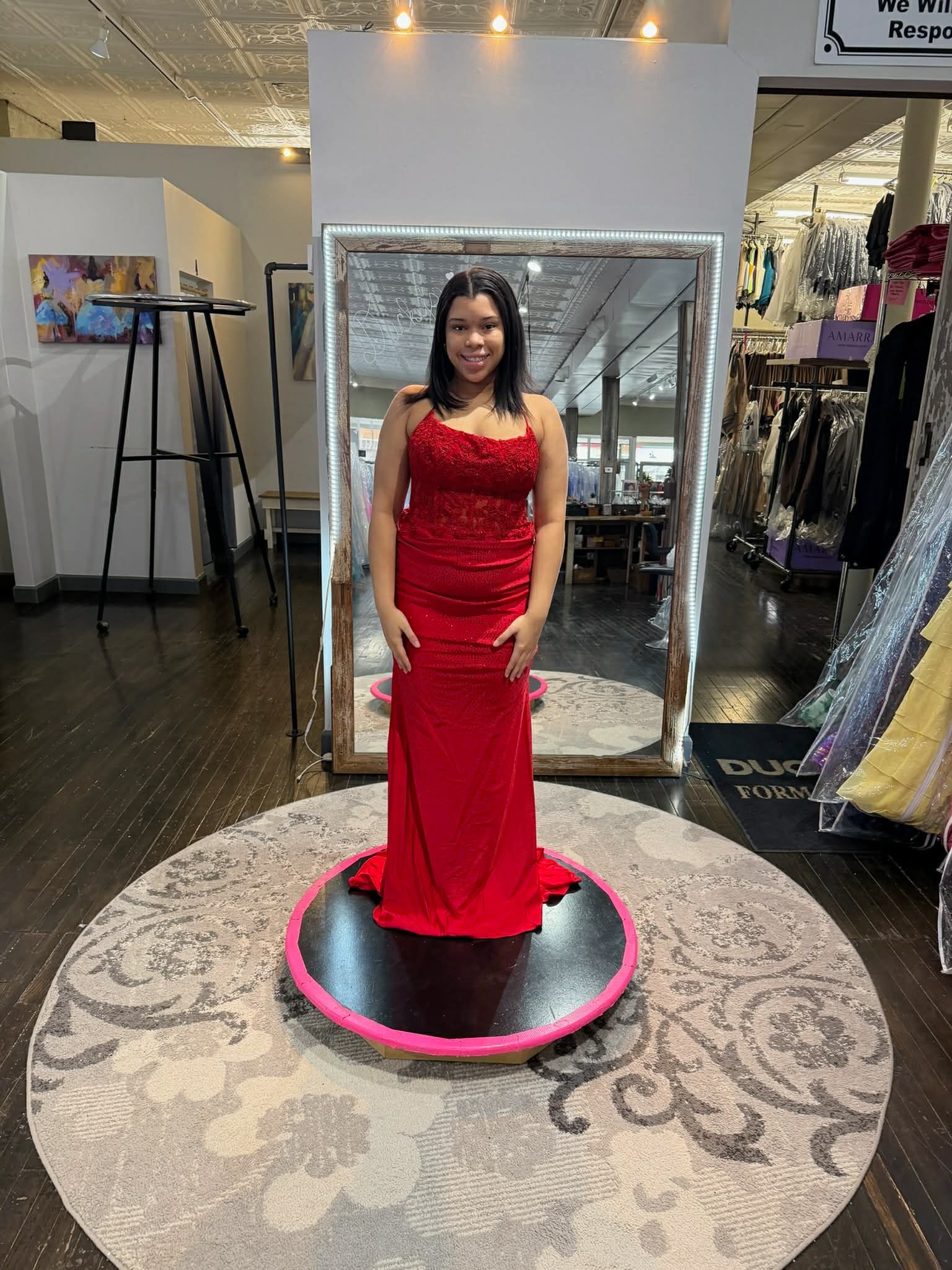 A person stands on a rotating platform in a clothing store, wearing a floor-length, strapless, red beaded evening gown.