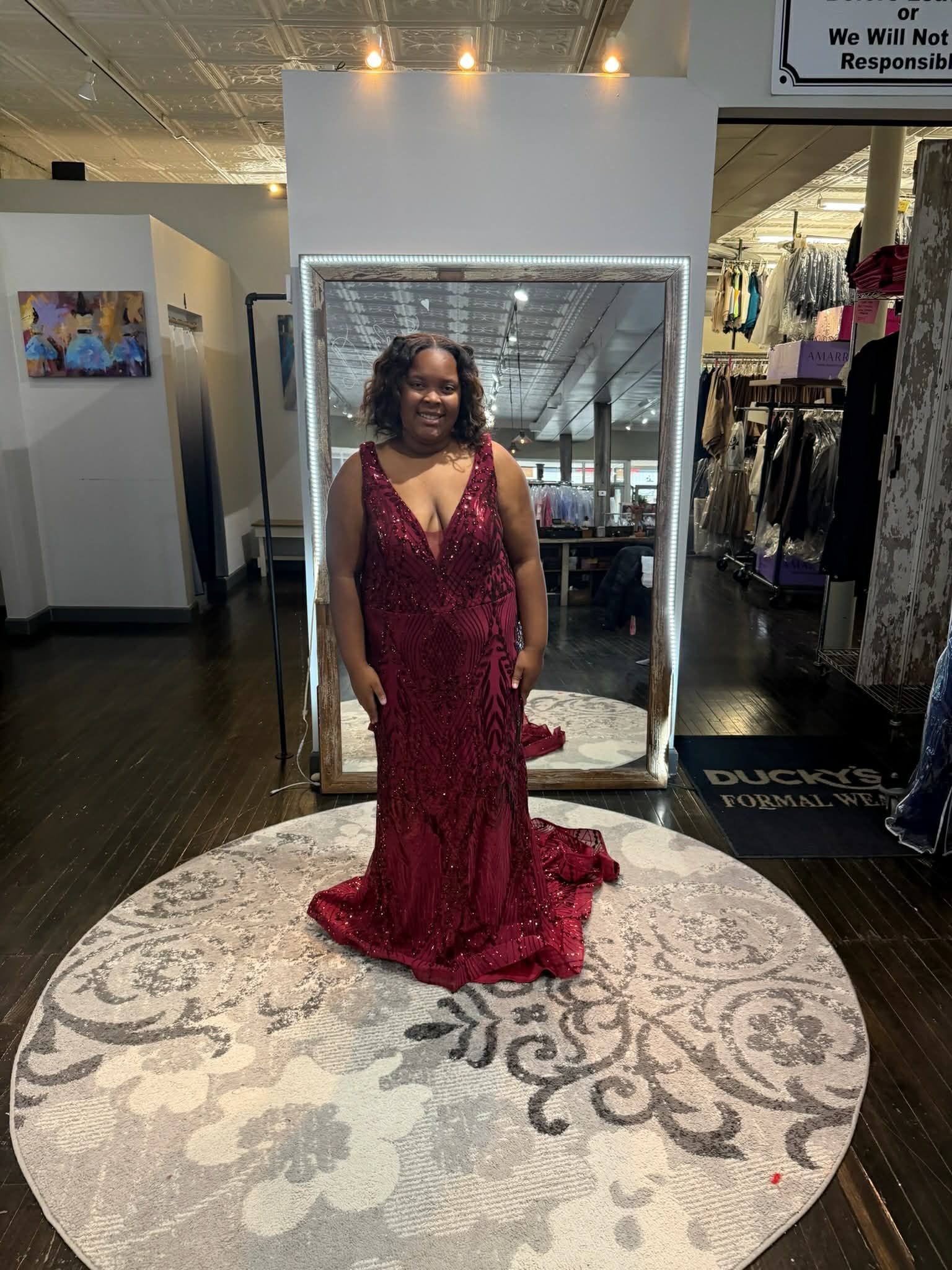 A person wearing a sparkling, deep red floor-length gown stands in a clothing boutique in front of a large mirror.