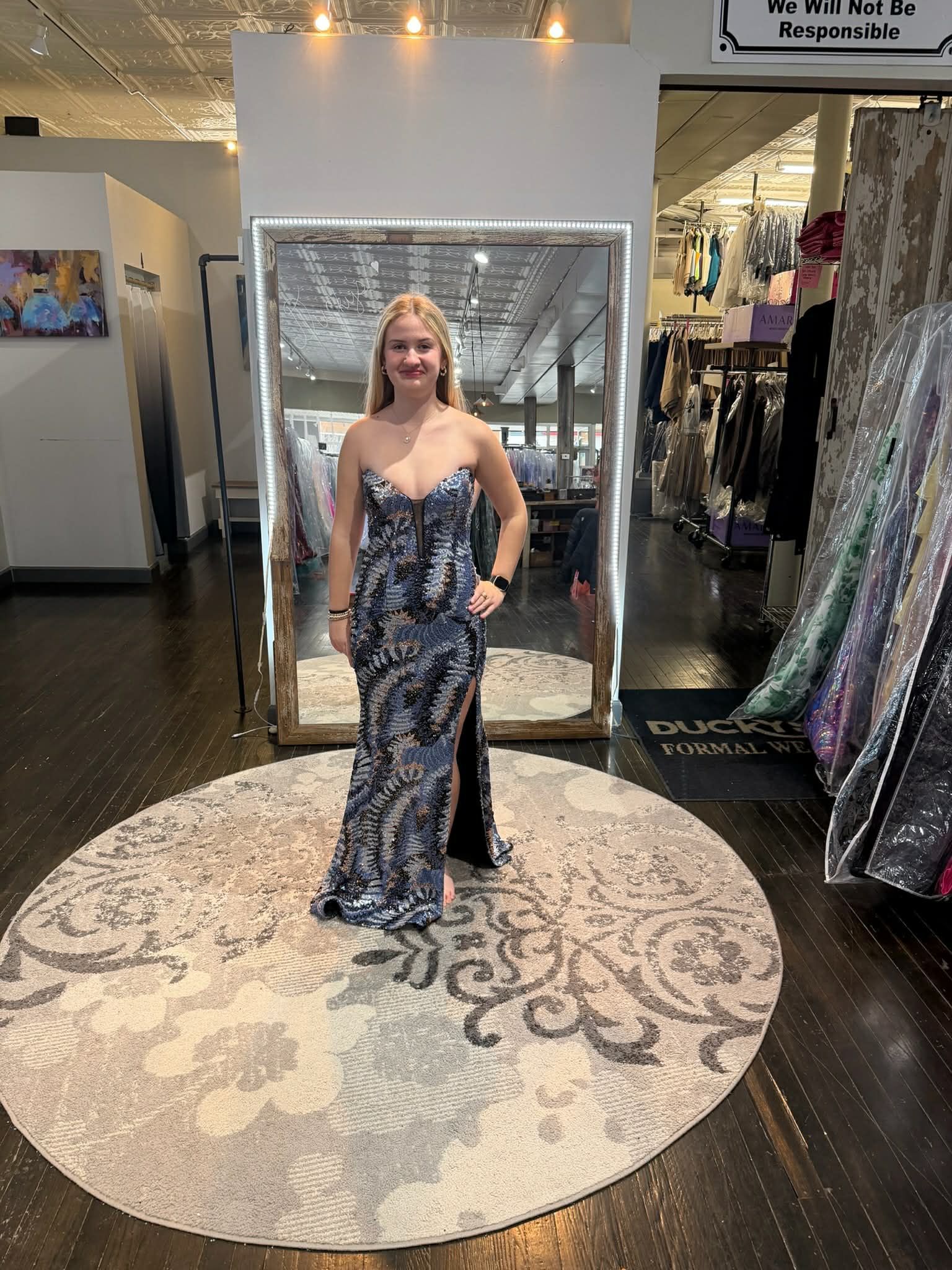 A person stands in a dress shop wearing a floor-length, dark, patterned strapless gown with a high leg slit.