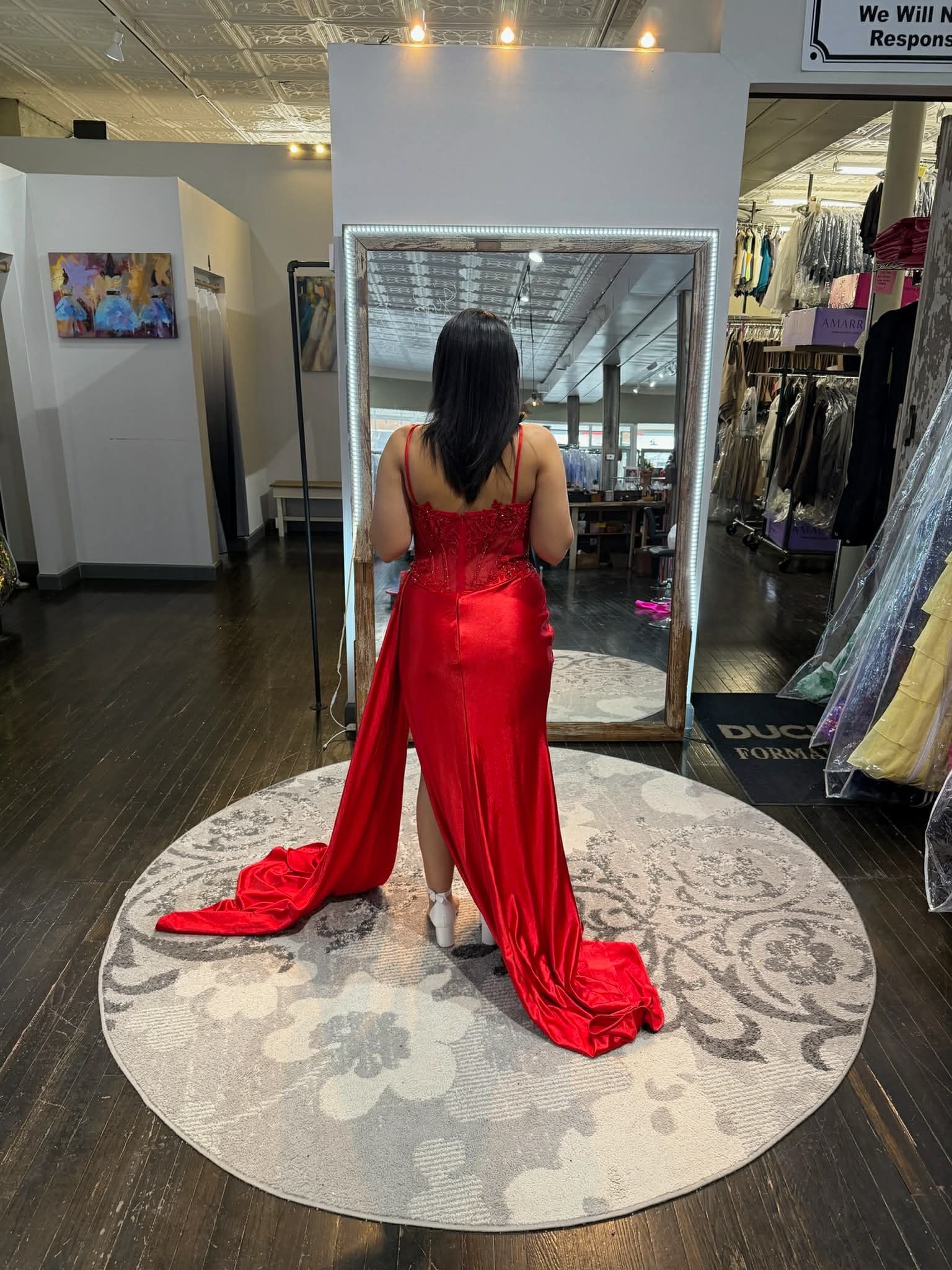 A person stands in a clothing boutique wearing a floor-length red satin dress with a train, facing a large mirror.