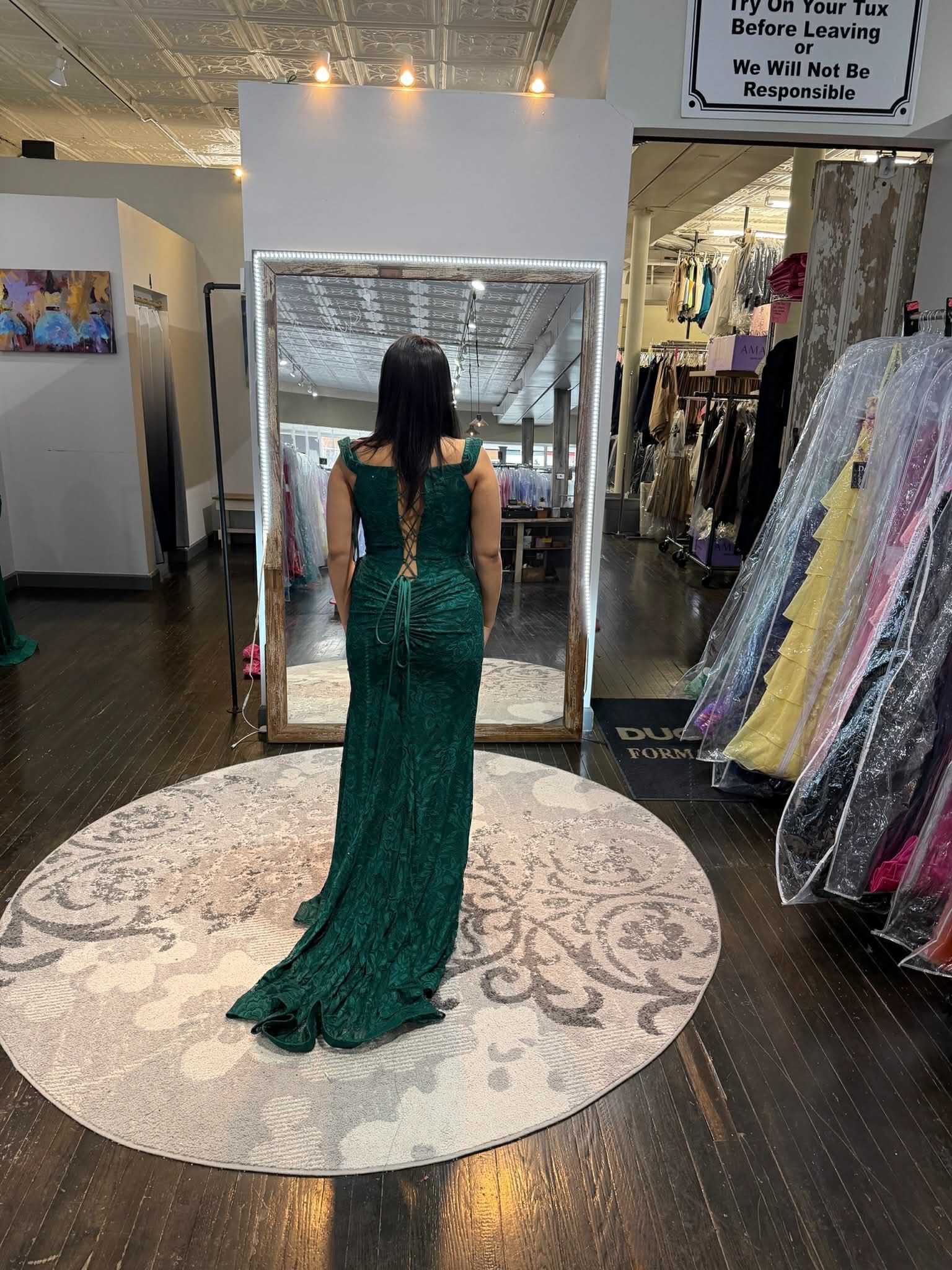 A person wearing a dark green, floor-length lace gown with a train, standing on a circular rug in a formal wear boutique.