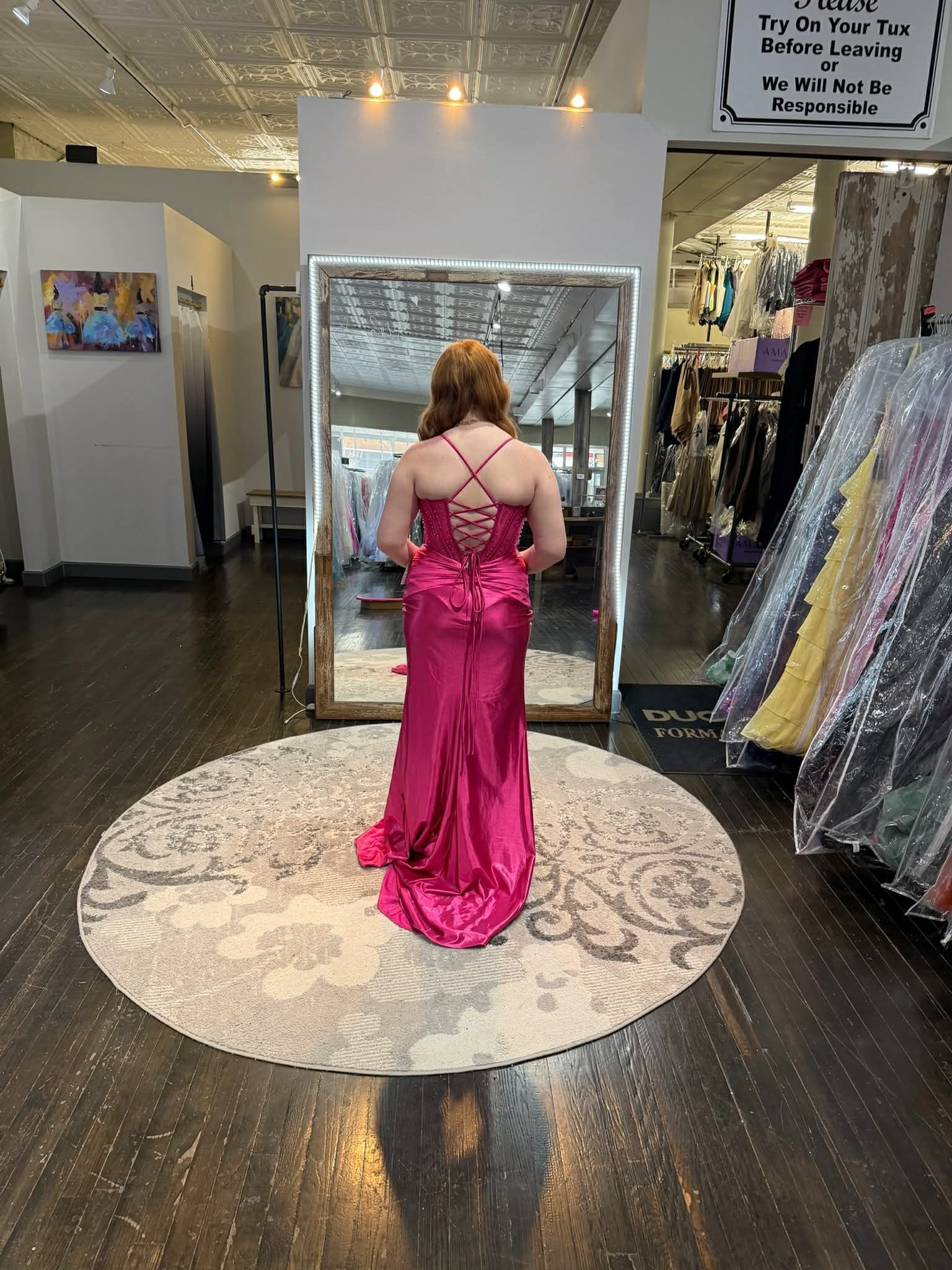 A person stands in a dress shop wearing a floor-length, magenta satin gown with a corset back, facing a large mirror.