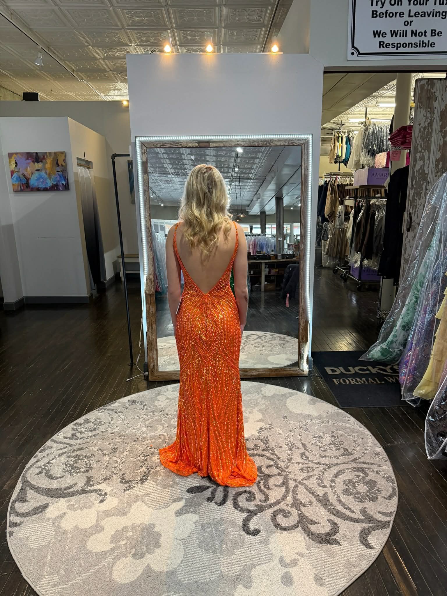A person stands in a dress shop wearing a floor-length, backless orange sequined gown, viewed from behind in a mirror.