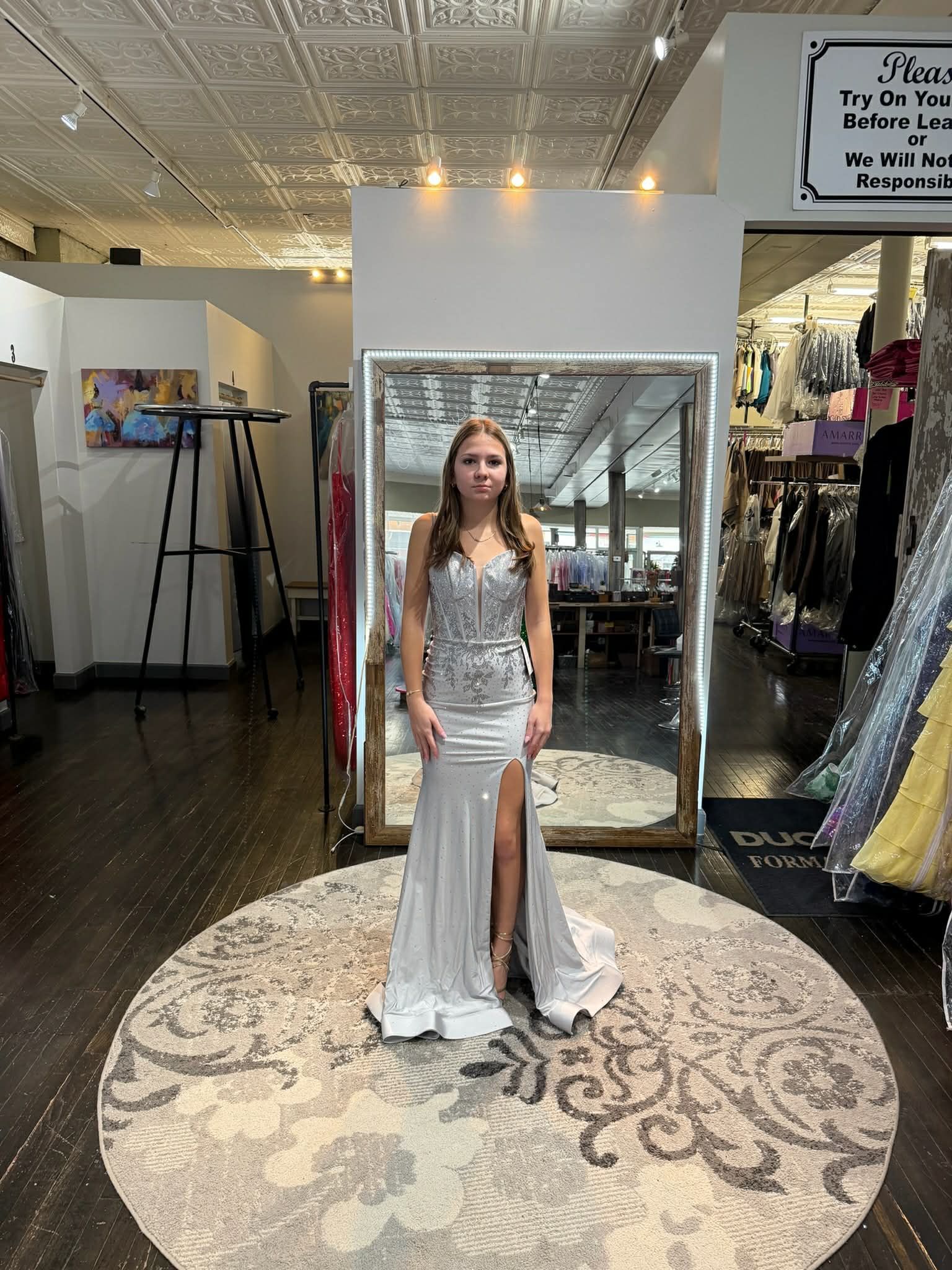 A person stands in a dress shop wearing a floor-length, silver, sequined gown with a high slit, posing before a mirror.