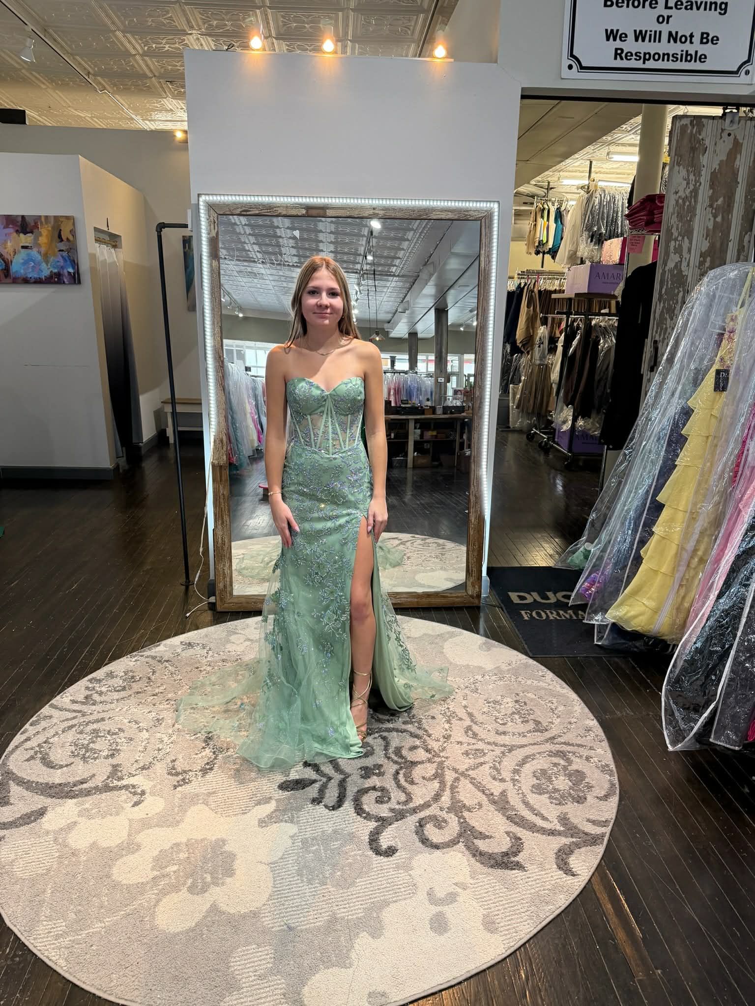 A person stands in a dress shop wearing a sage green, embellished, strapless floor-length gown with a high side slit.