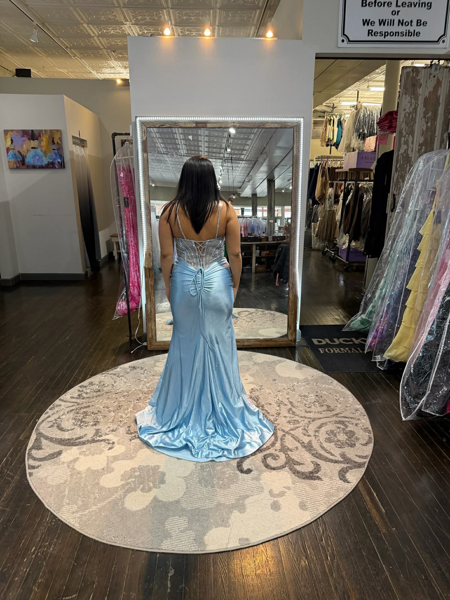 A person stands in a dress shop, facing a mirror while wearing a light blue, floor-length gown with a corset-style back.