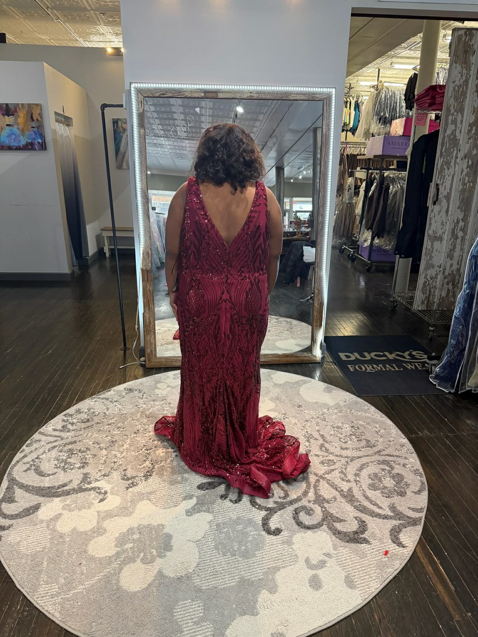 A person wears a floor-length, deep red sequined gown with a V-back and short train, standing before a large mirror.